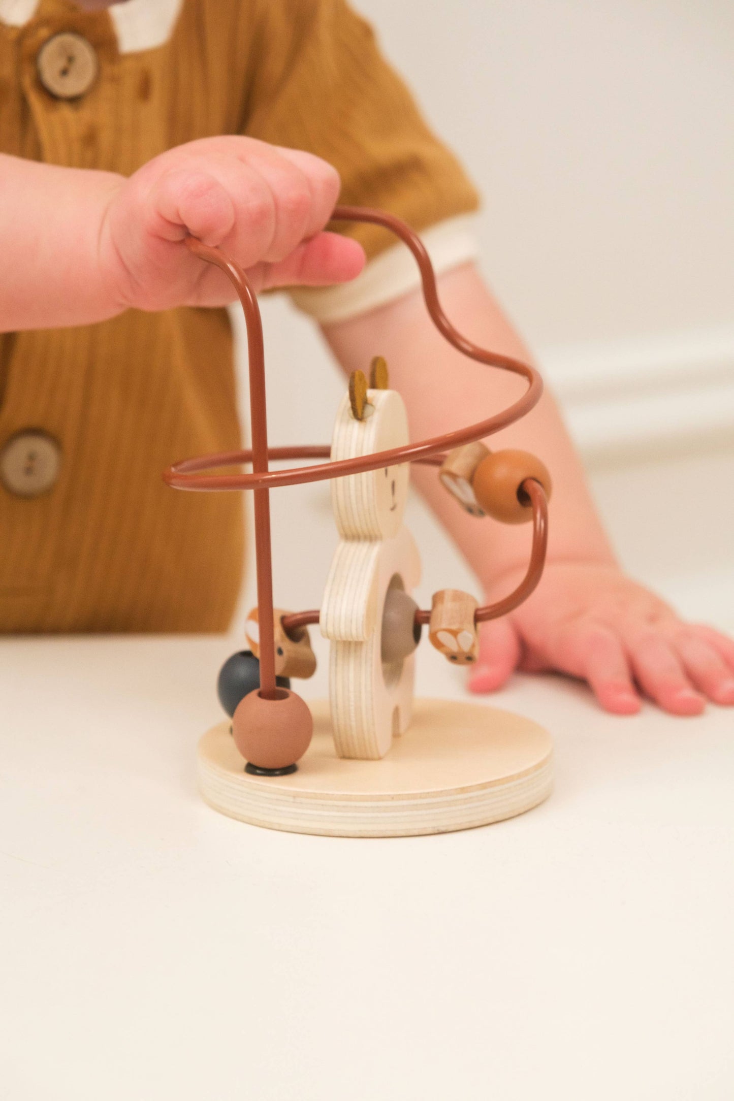 Wooden toy with beads on a stand held by a hand, with a blurred background of a person wearing a mustard yellow shirt.