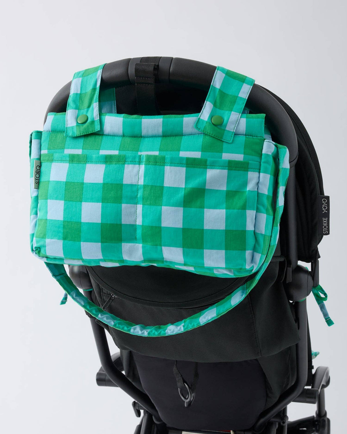 A green and white gingham baby diaper bag with top handles and a matching shoulder strap, mounted on a black stroller against a light gray background.
