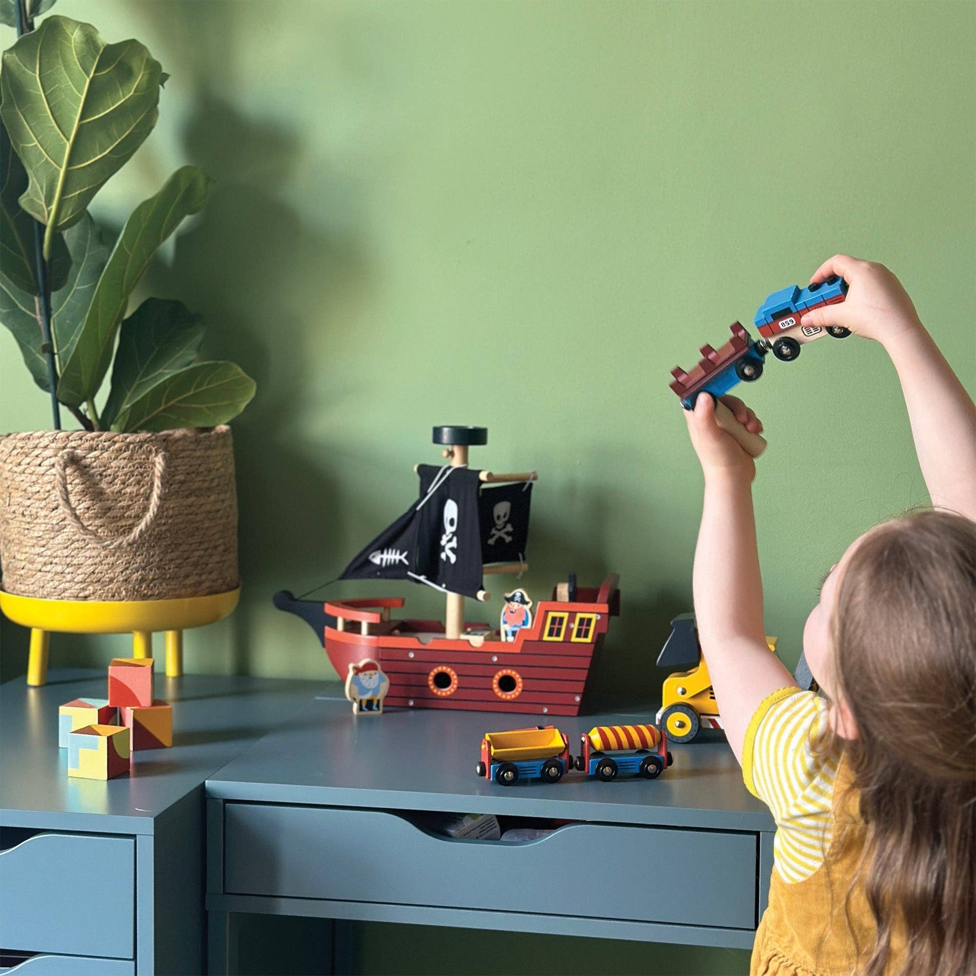 A child holding a wooden train set with one engine and three colorful attached carriages designed to resemble industrial cargo goods up in the air against a green background.