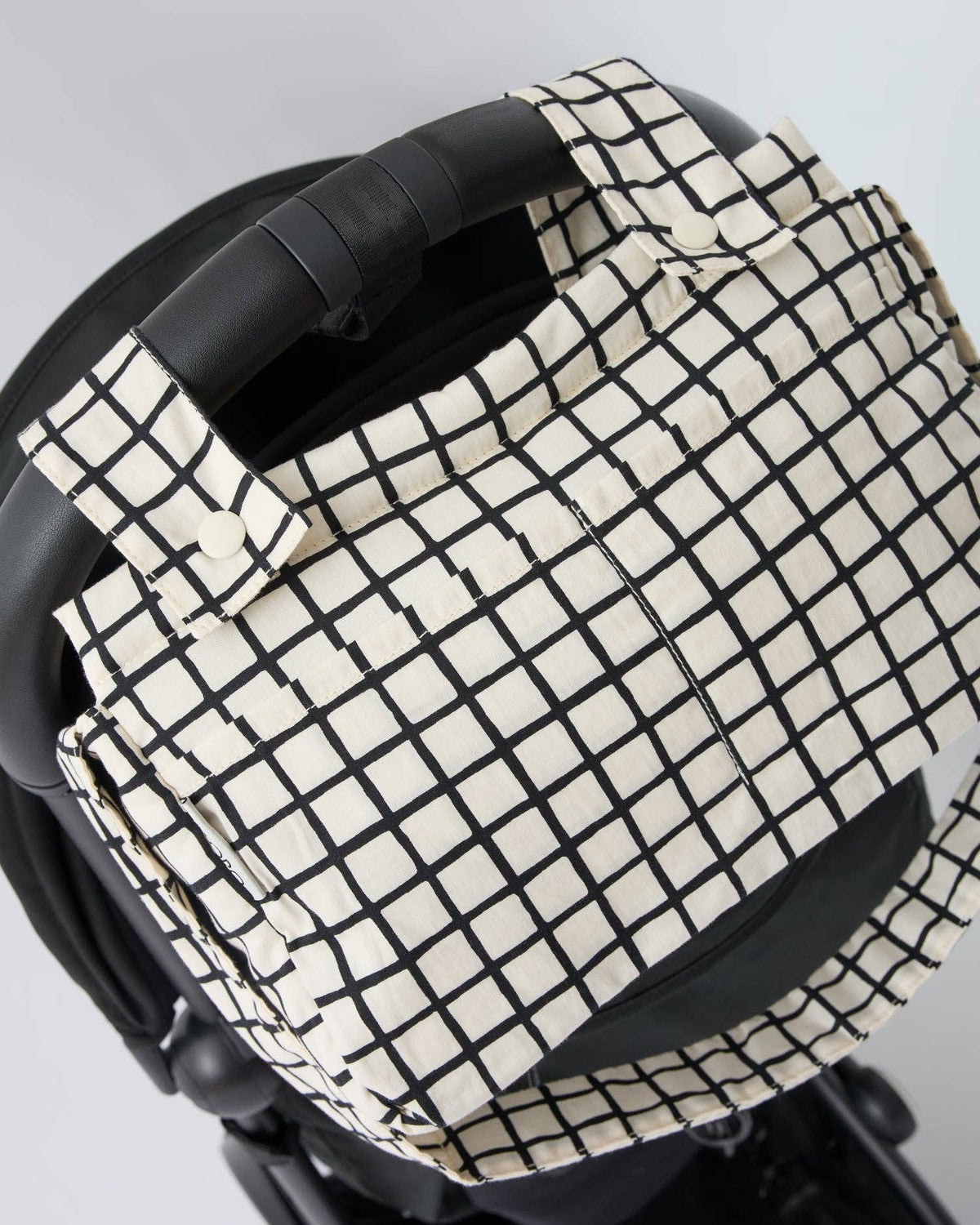 A close-up of a black and off-white grid patterned bag or cover attached to a black stroller or pram handle, shown against a light gray background.