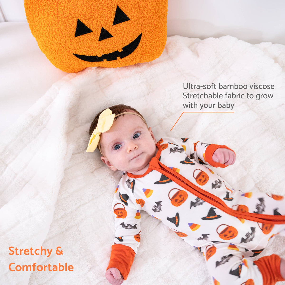 A baby lying on a white quilted blanket wearing a white one-piece pajama with orange trim and a pumpkin, bat, and candy corn print. A yellow bow headband is also visible.