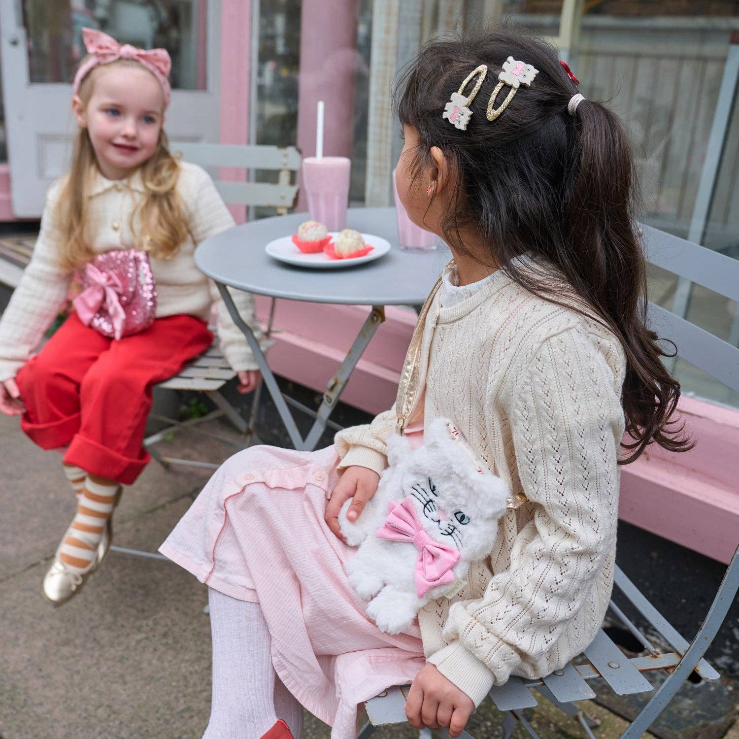 Two young girls sitting outdoors at a small table with a pink drink and cake, wearing matching outfits and hair accessories.
