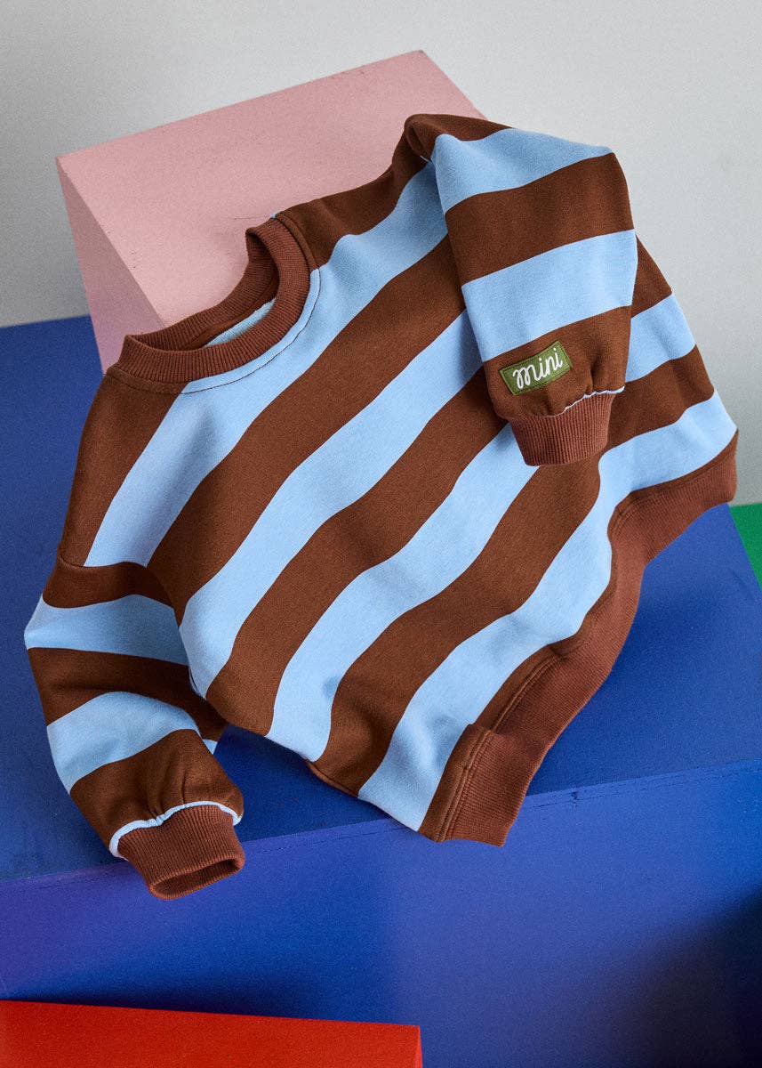 A long-sleeve crewneck sweatshirt with alternating brown and light blue horizontal stripes, styled on a colorful geometric block background. A small green 'mufe' brand tag is visible on the left chest pocket.