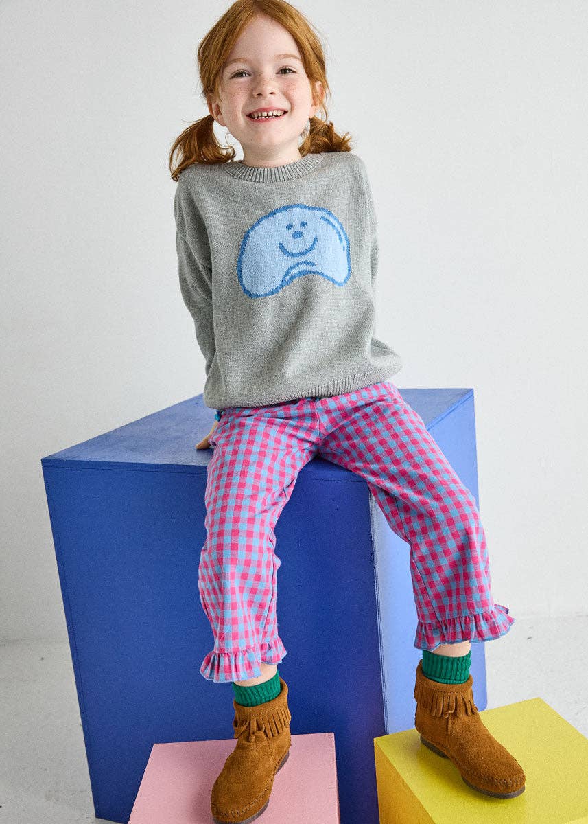 A young girl with red hair in pigtails sits on a blue block, wearing a gray knit sweater with a blue smiley face graphic, pink and white gingham pants, green socks, and brown suede boots.