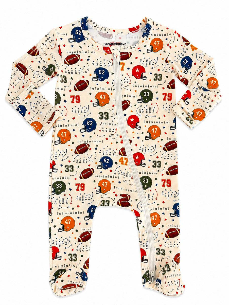 A baby long-sleeve footed sleeper with a front zipper, featuring an all-over football-themed print on a cream background with helmets, footballs, numbers, and stars.