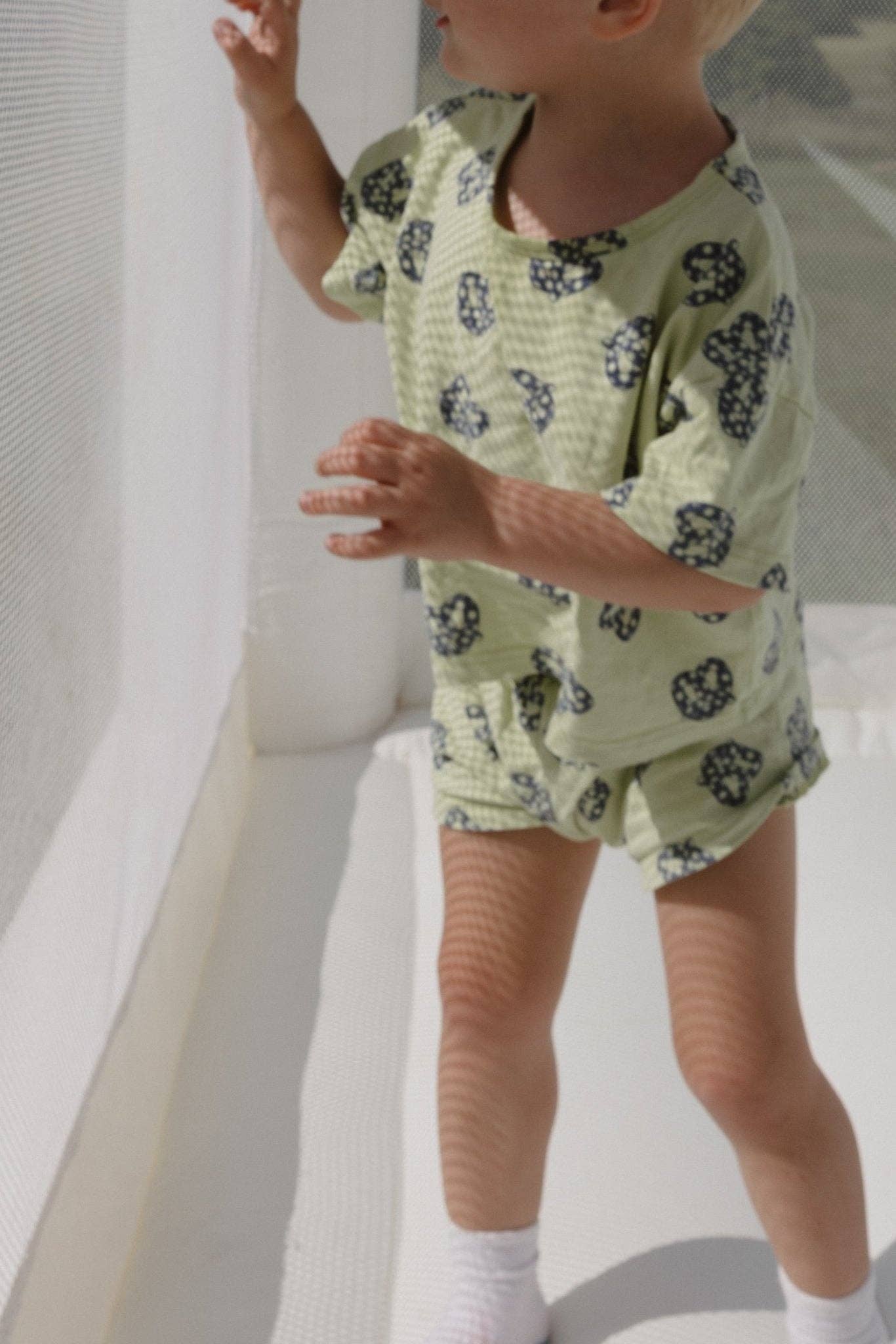 Child wearing a light green outfit with blue pretzel patterns in a minimal indoor setting.