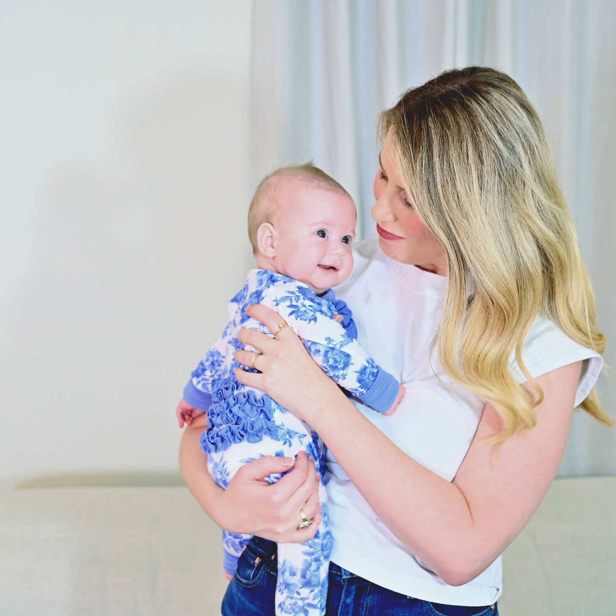 A woman holding a baby wearing a blue and white floral patterned romper, set against a soft neutral indoor background.