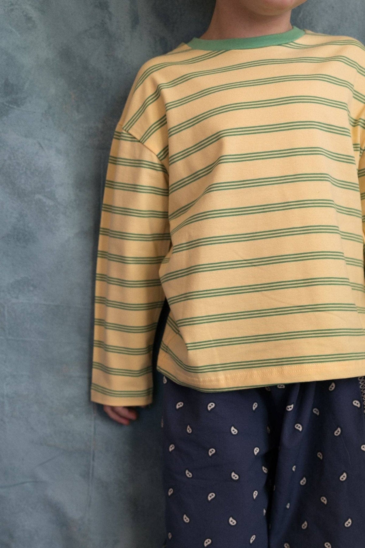 A child wearing a long-sleeve yellow shirt with thin green horizontal stripes and a green crew neck, paired with navy blue paisley pants, standing against a textured gray wall.