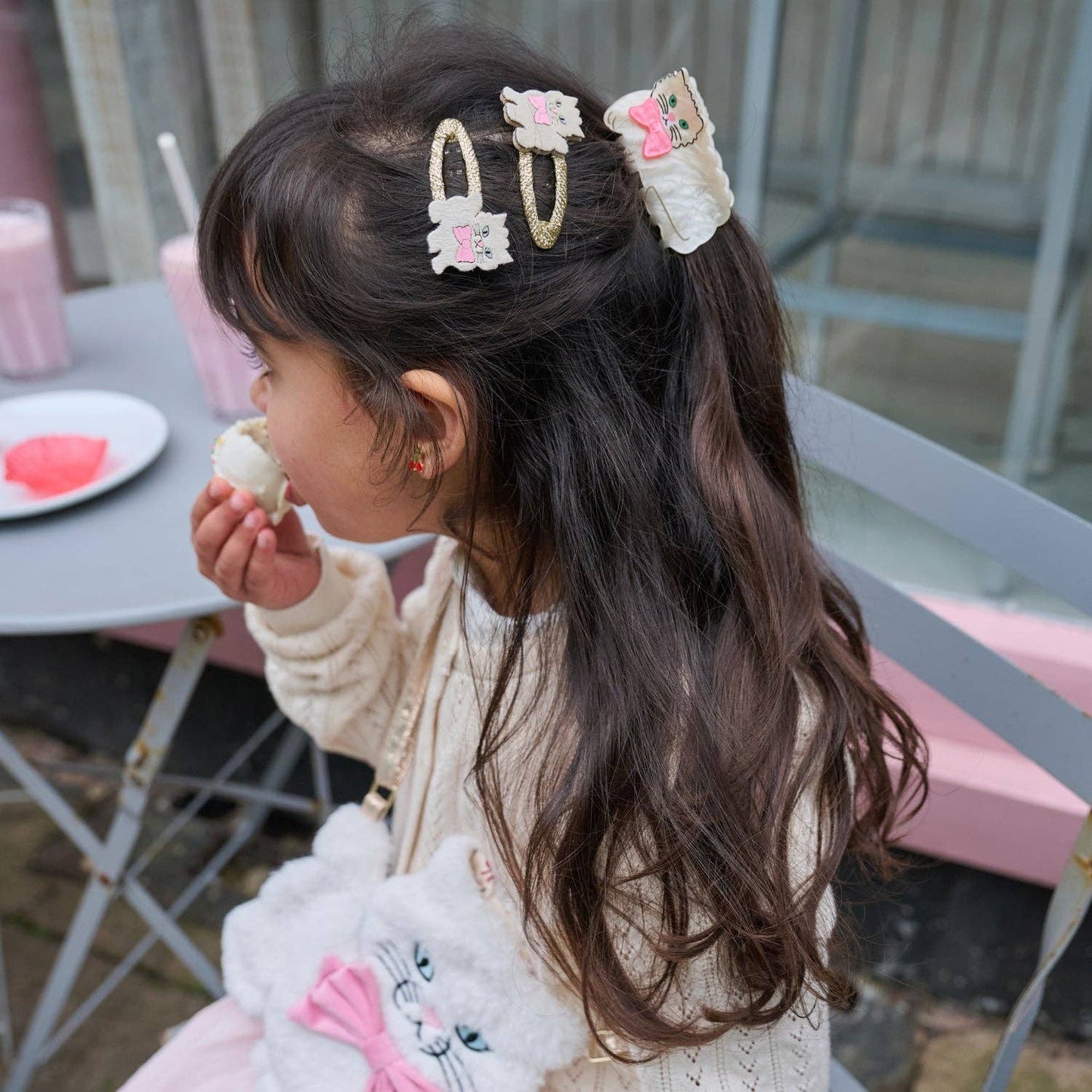 A young girl with long dark brown hair wearing three cute hair clips, sitting at a pink table and eating a cookie. The hair clips feature pastel pink, white, and gold designs.