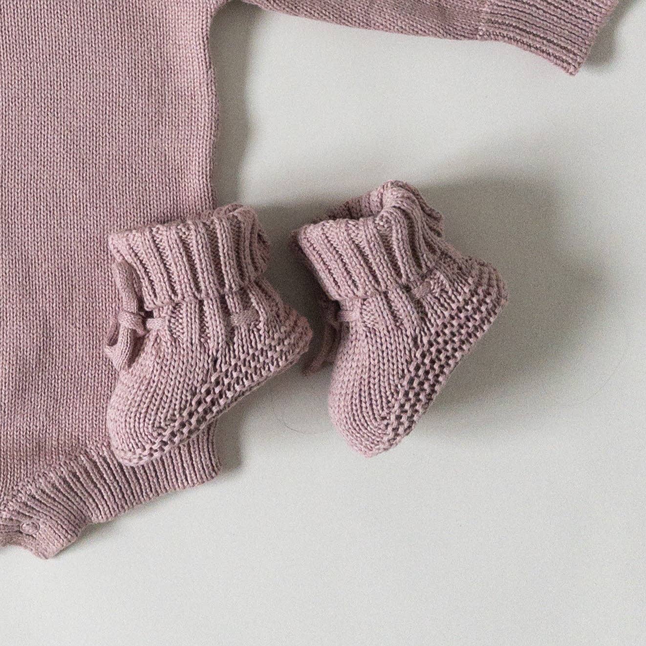 A pair of dusty pink hand-knit baby booties with ruffled cuffs and small bows, displayed next to a matching knit baby sweater on a light gray background.
