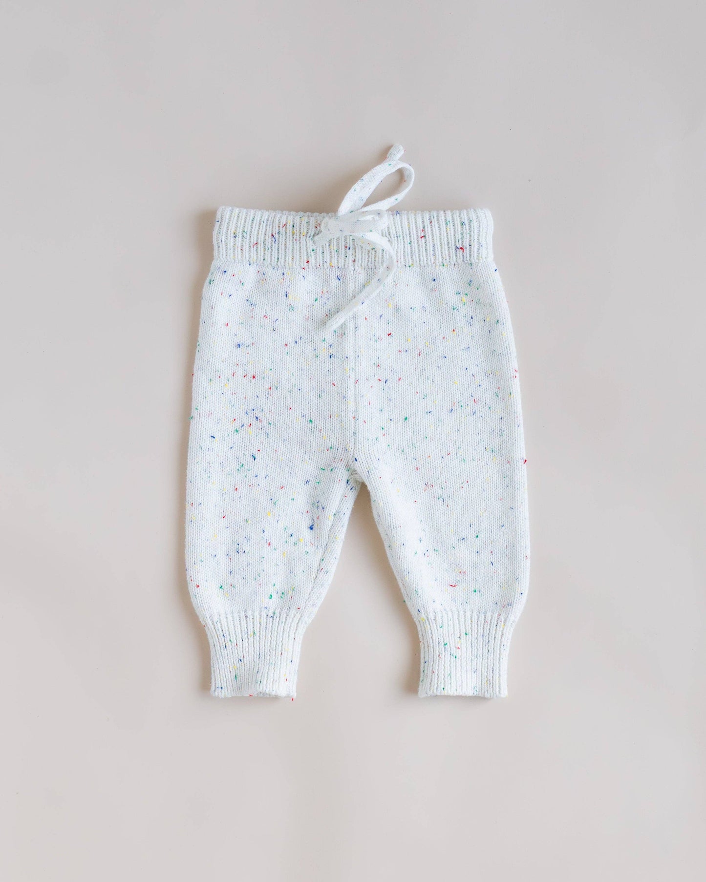 White knit baby pants with a colorful confetti speckle pattern, featuring ribbed cuffs and a drawstring waist, laid flat on a light beige background.