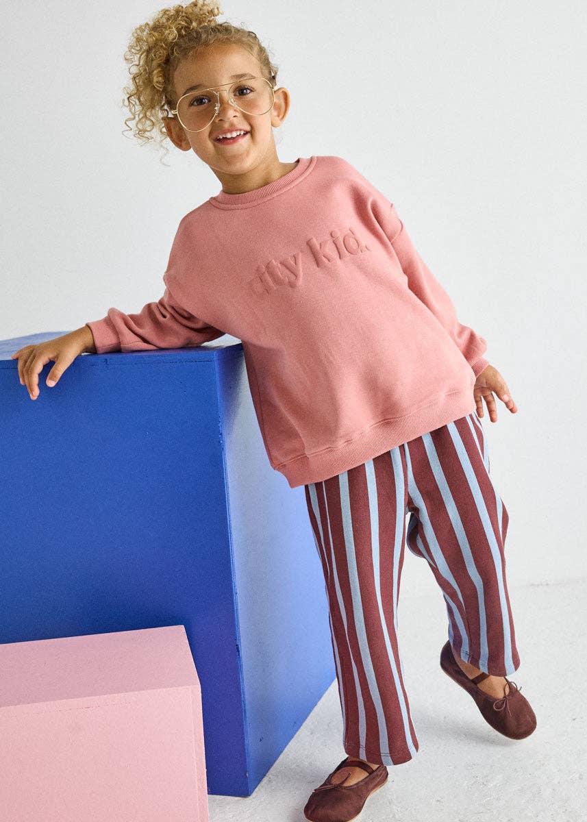 A young girl with curly hair and glasses is smiling while posing in a pink crewneck sweatshirt with 'city kid' embroidered on the chest, paired with red and white vertically striped pants and brown shoes. She is leaning against a blue block next to a pink block on a white studio background.