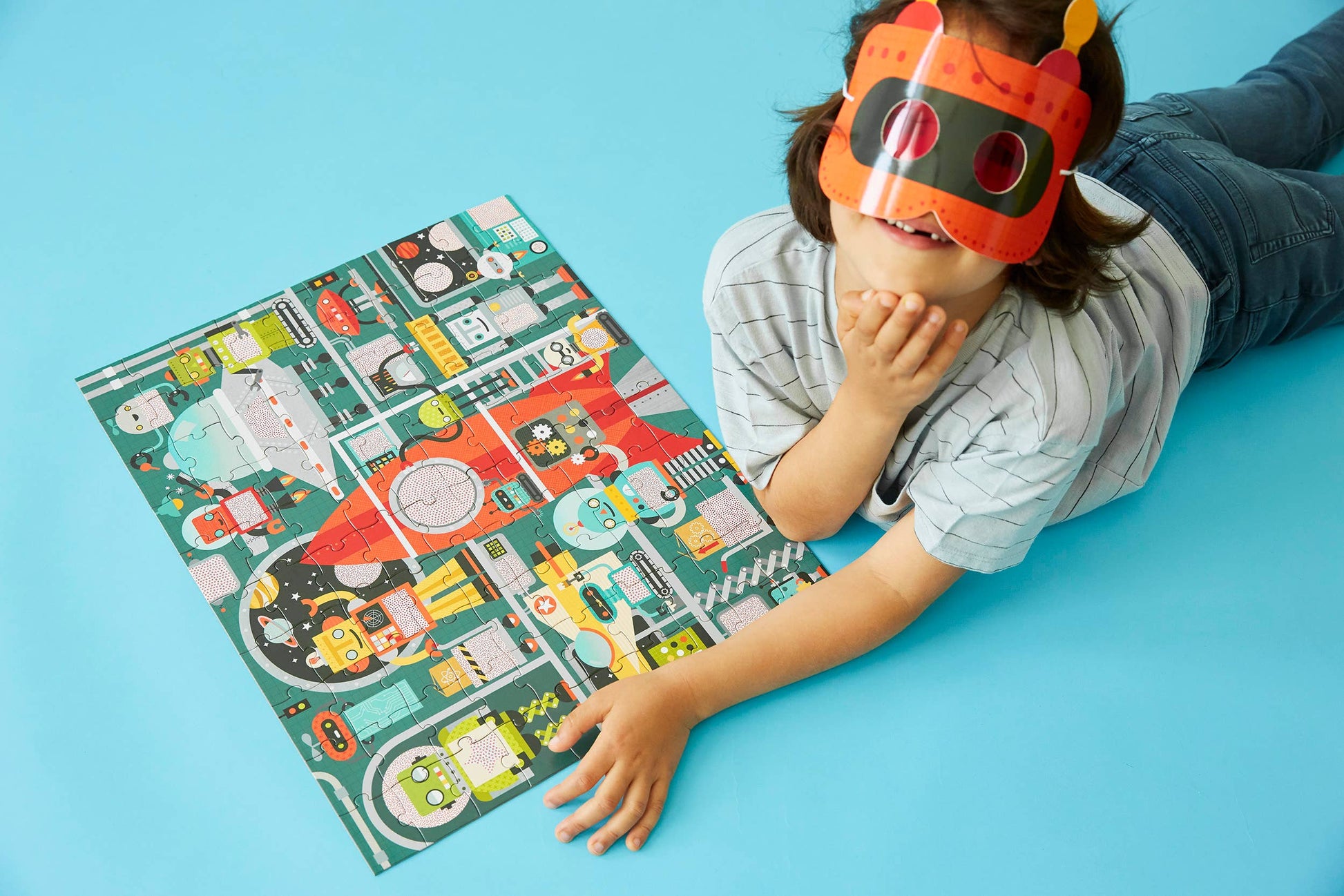 Child wearing a robot mask lying on a blue surface with a colorful robot-themed puzzle.