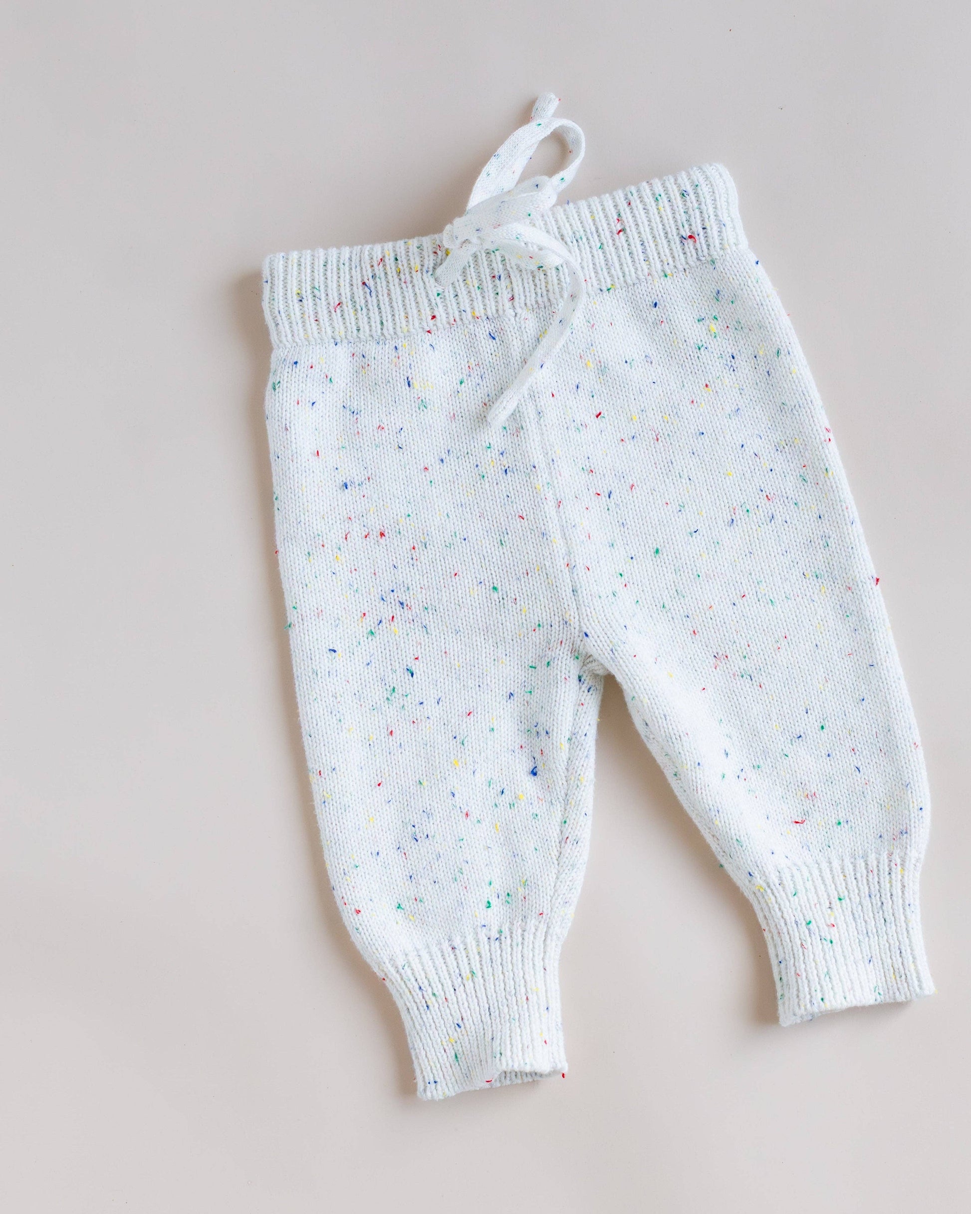 White knit baby pants with a multicolor speckled pattern, featuring a drawstring waist and ribbed cuffs, laid flat on a light beige background.