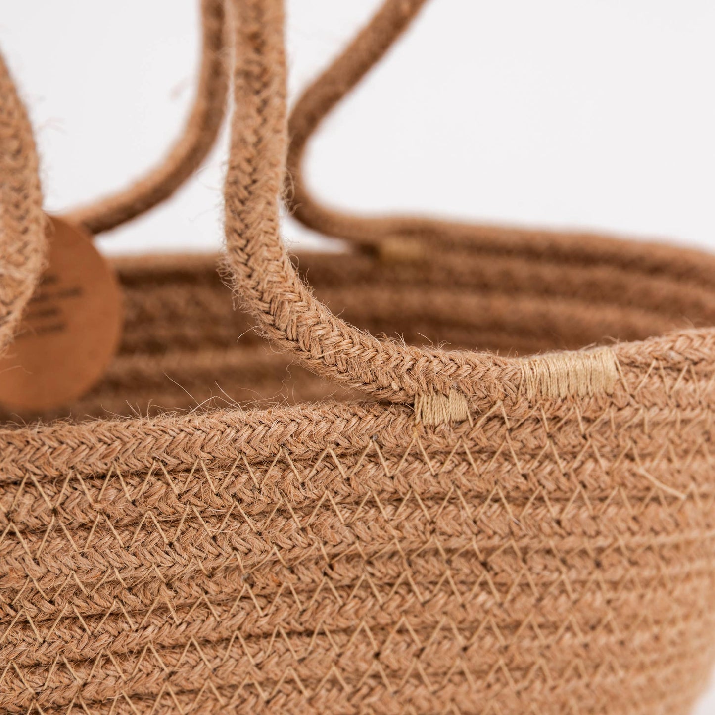 Close-up of a woven natural fiber basket in a warm tan-brown color, showing the braided texture and part of a round wooden tag on a white background.