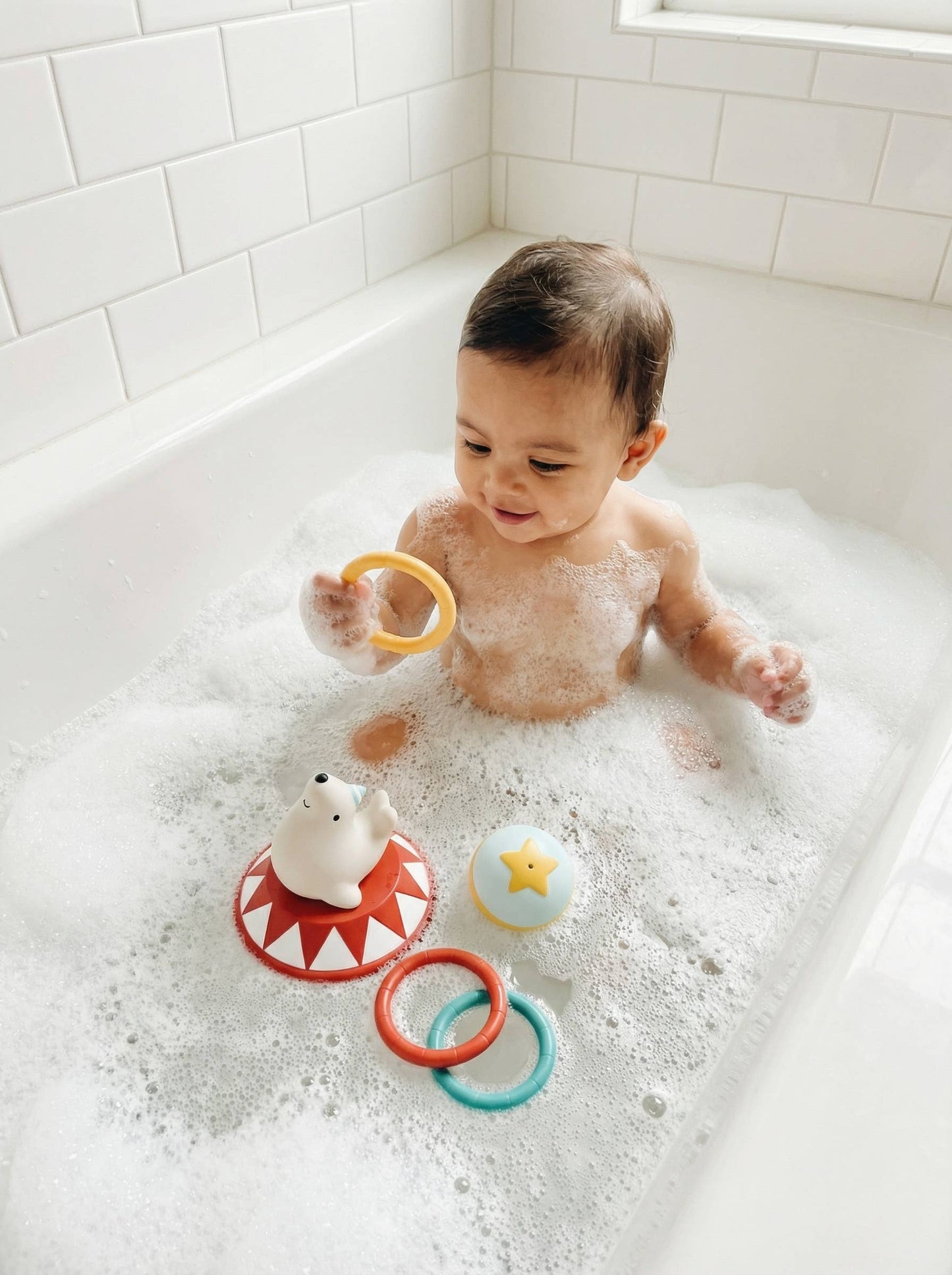 A baby sitting in a bubble-filled bathtub playing with colorful bath toys, including a yellow ring, a red and white seal toy, and a blue star toy.