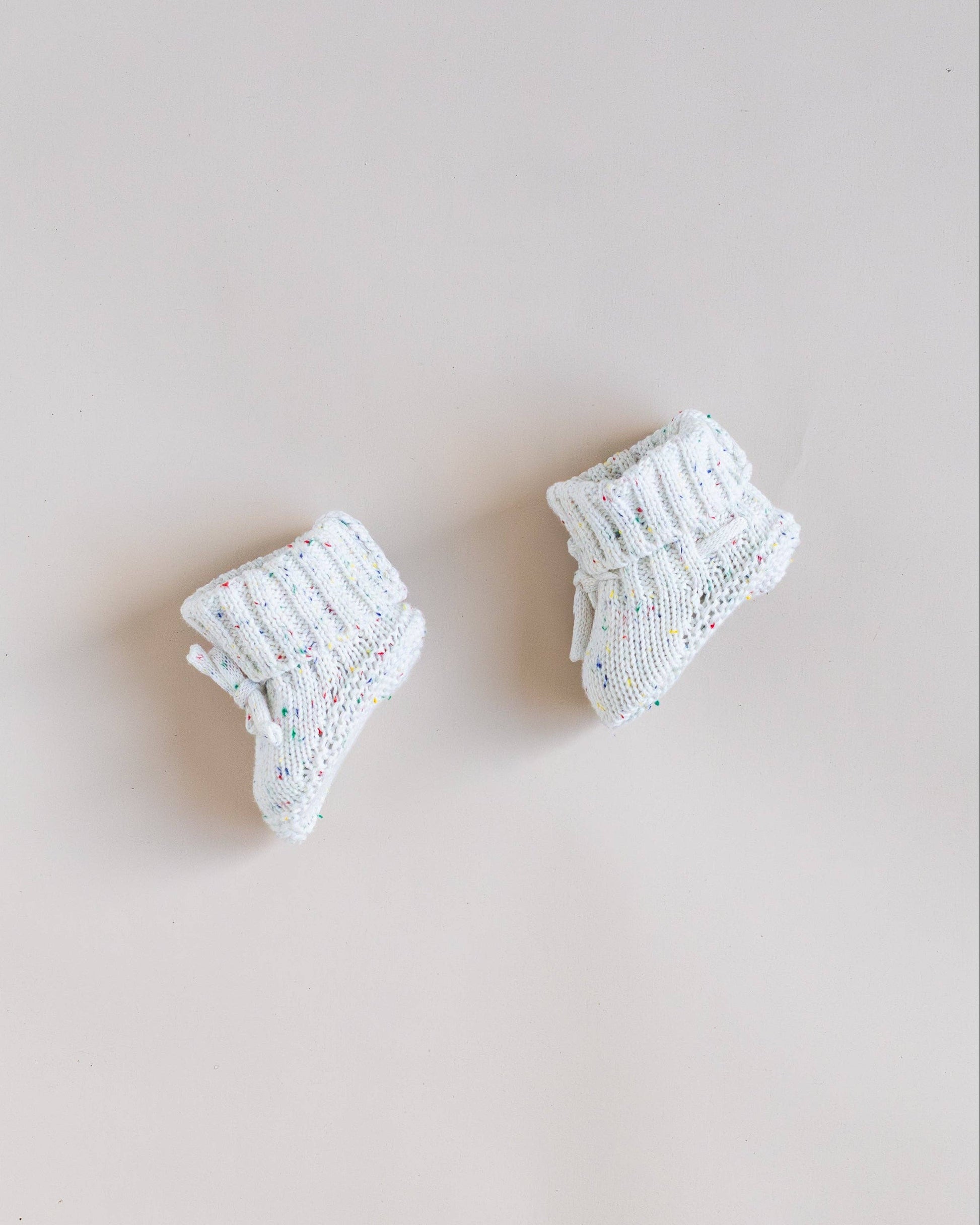 Pair of white baby booties with colorful speckles on a light beige background
