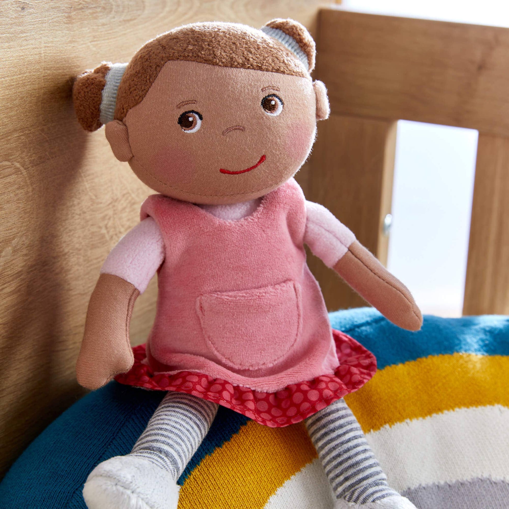 Plush toy doll with a pink dress sitting on a colorful rug.