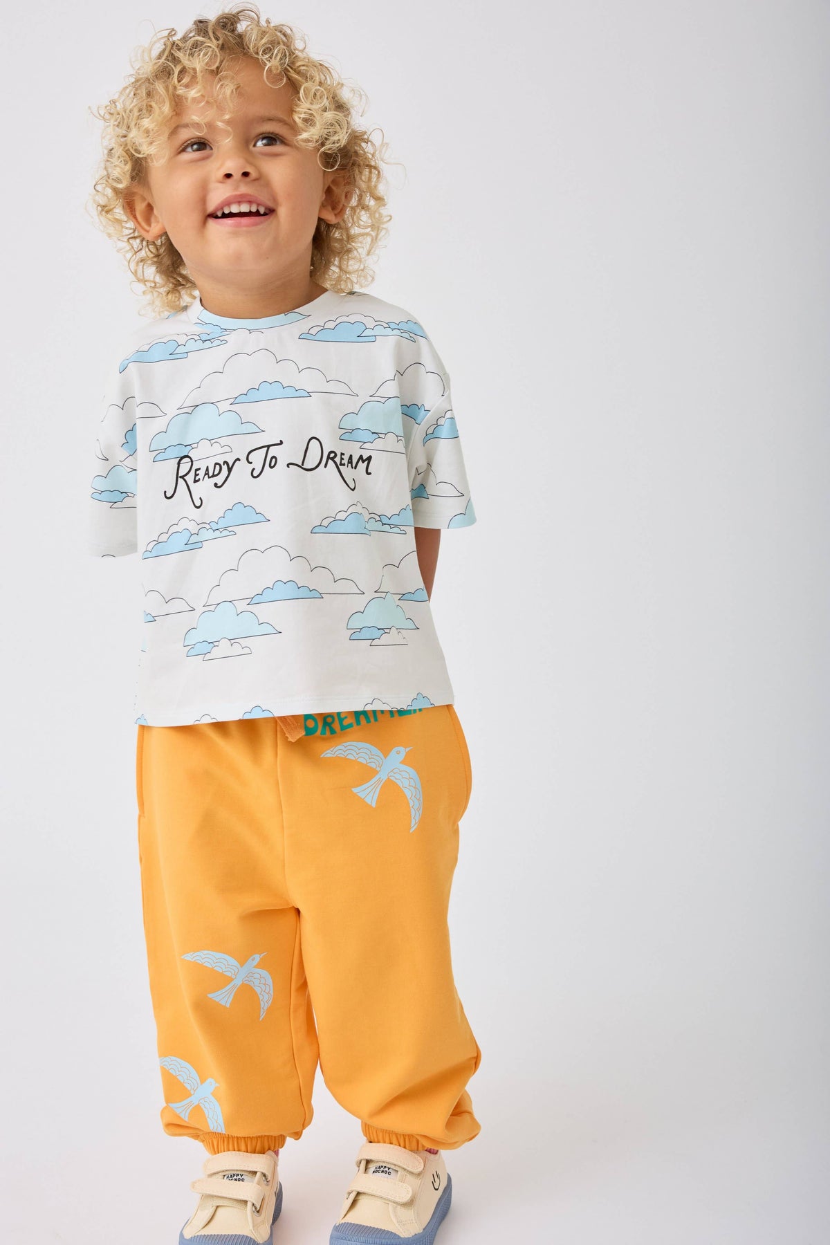 A toddler with curly blonde hair stands against a light gray studio background wearing a white graphic T-shirt with blue clouds and the words 'Ready To Dream' across the chest, paired with mustard yellow jogger pants featuring light blue bird prints.
