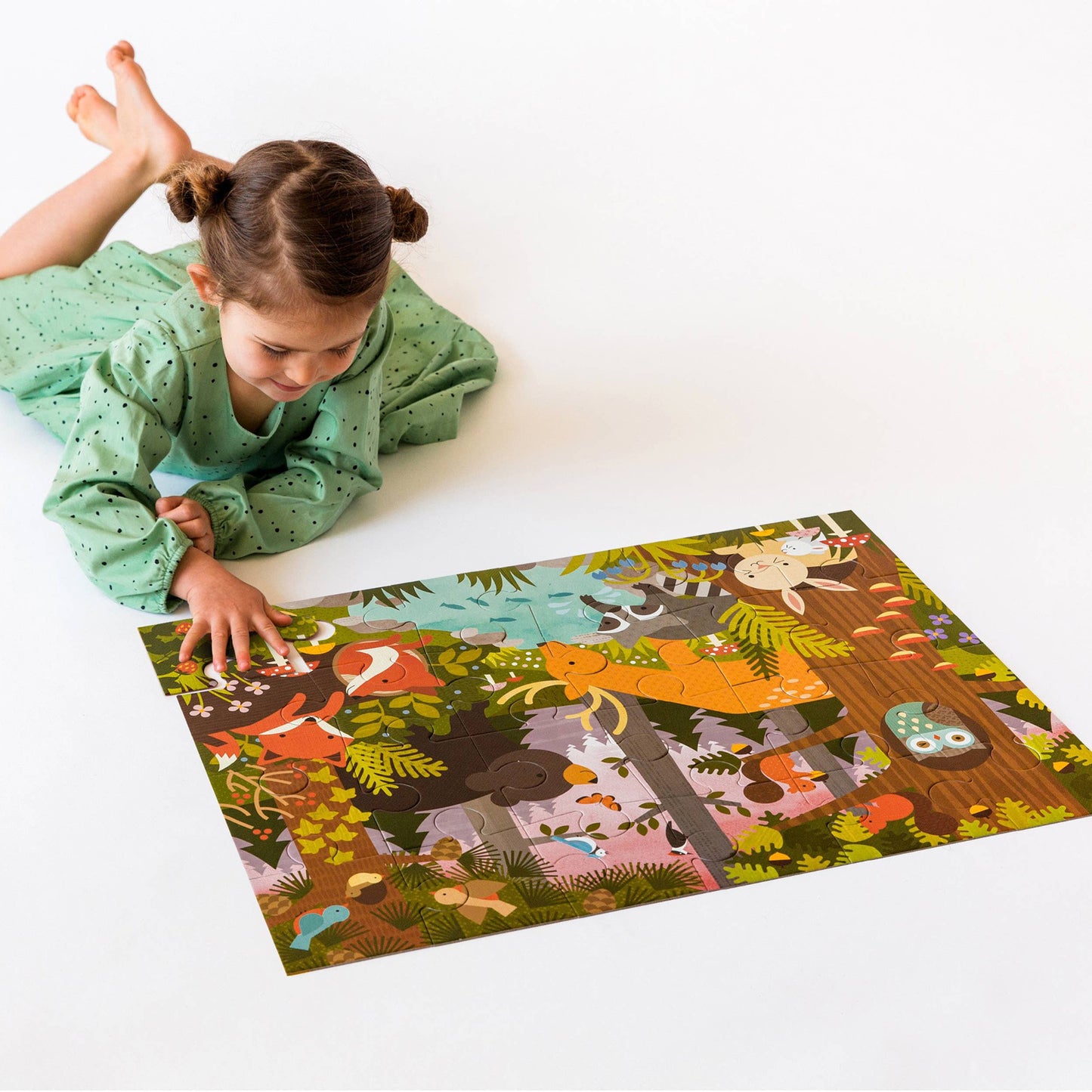 Child in green dress laying on floor with a 24-piece floor puzzle with illustrations of an enchanted woodland scene, including animals like deer, bears, foxes, and rabbits.