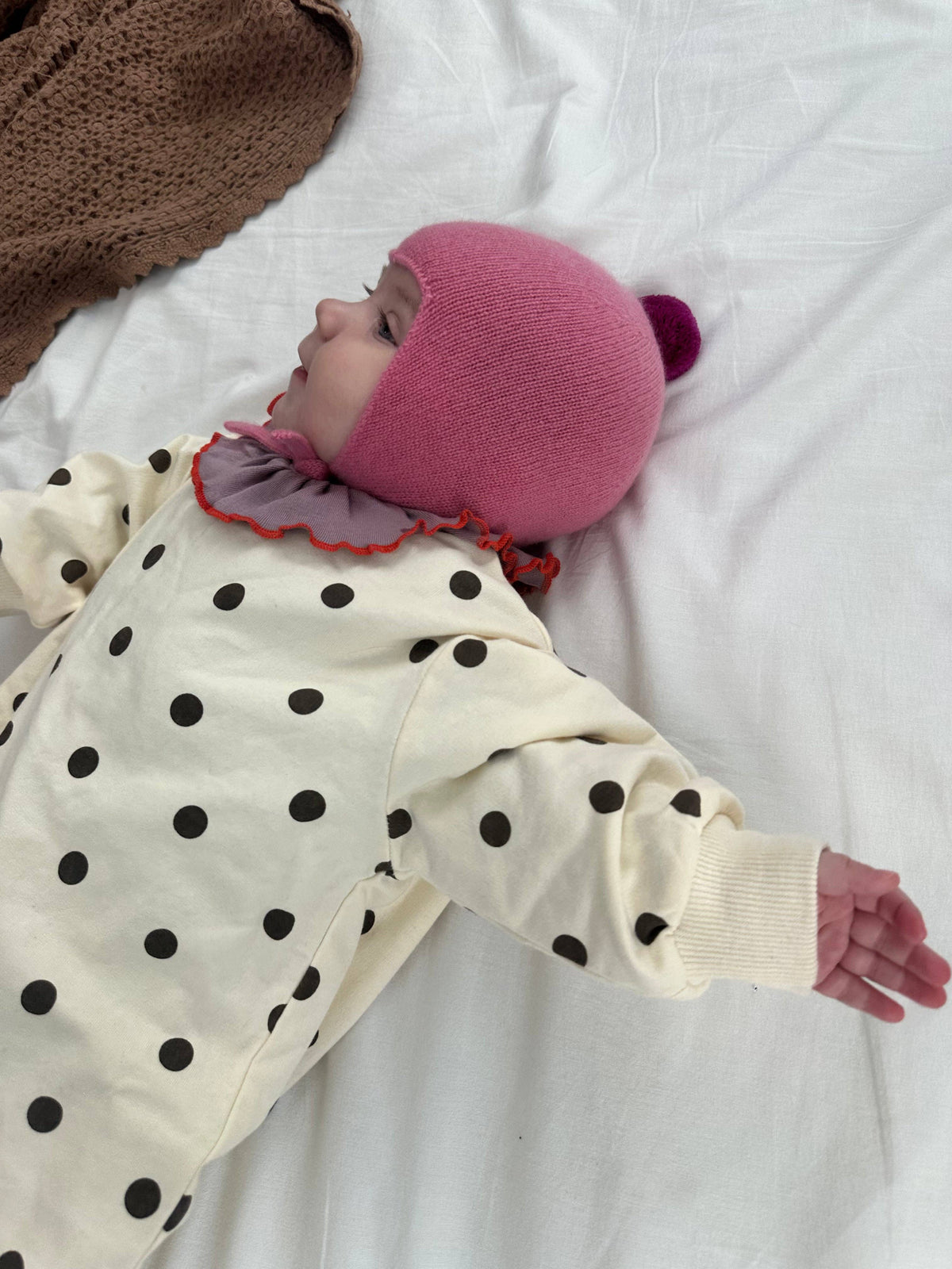 A baby lying on a white sheet, wearing a cream polka-dot onesie and a pink knit bonnet with a small pom-pom.