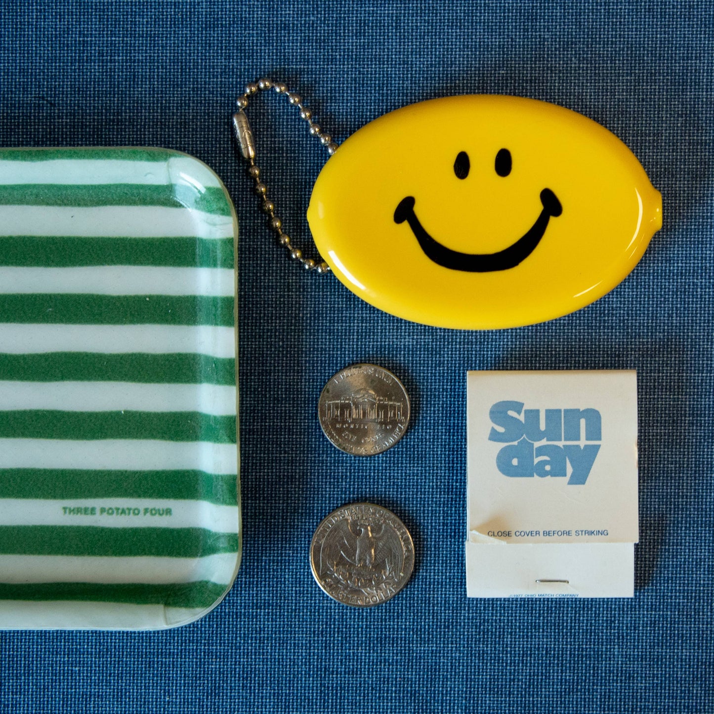 Green and white striped tray, yellow smiley face keychain coin purse, two coins, and a 'Sunday' product on a blue textured surface.