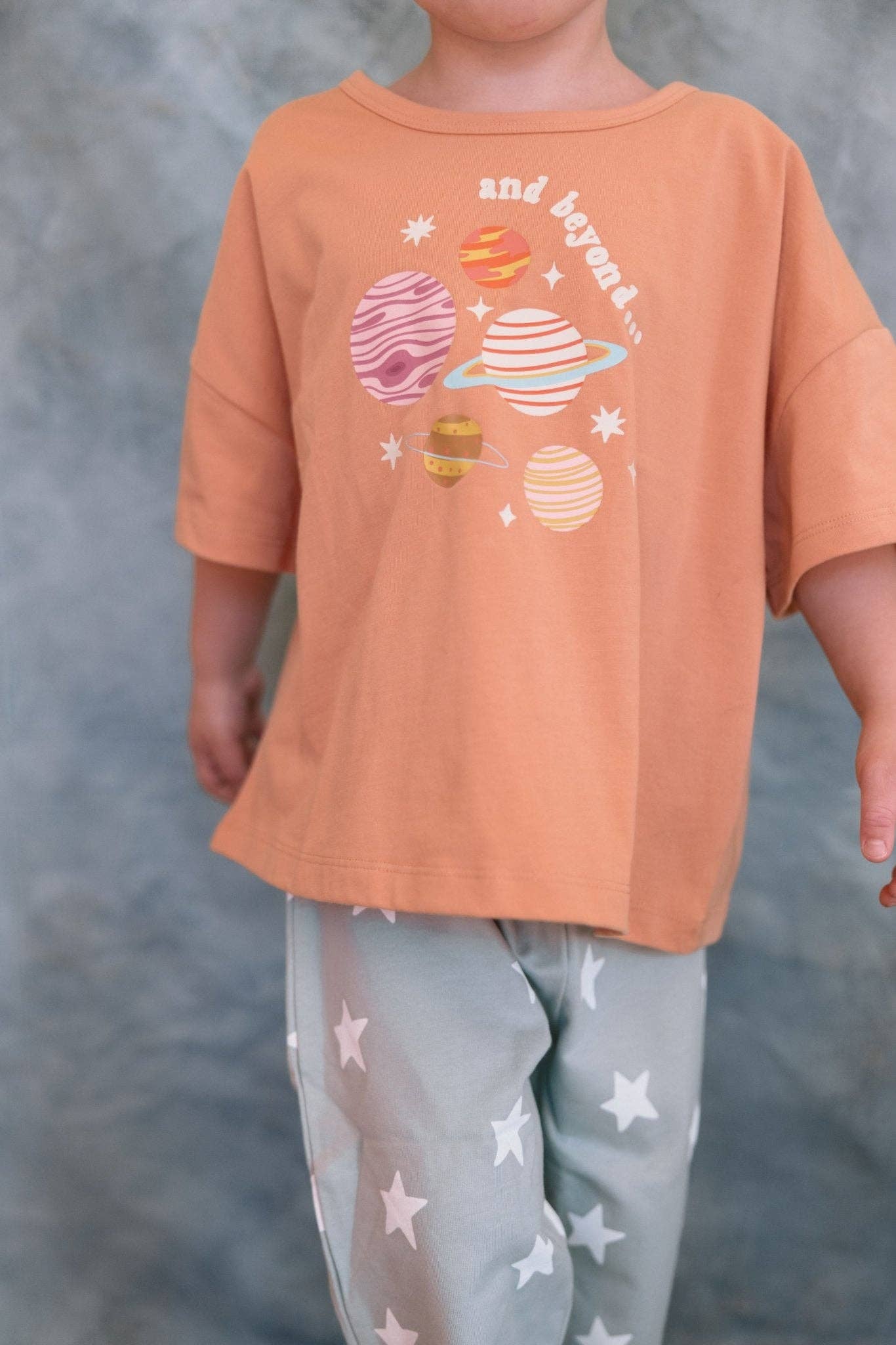 Child wearing an orange shirt with space-themed graphics and gray pants with star patterns against a gray background.