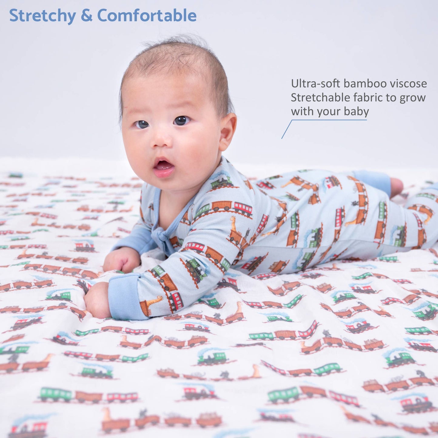 A baby lying on their stomach on a stretchy train-print blanket, wearing a light blue long-sleeve sleeper with a colorful train pattern. Text overlays read 'Stretchy & Comfortable' and 'Ultra-soft bamboo visco Stretchable fabric to grow with your baby'.
