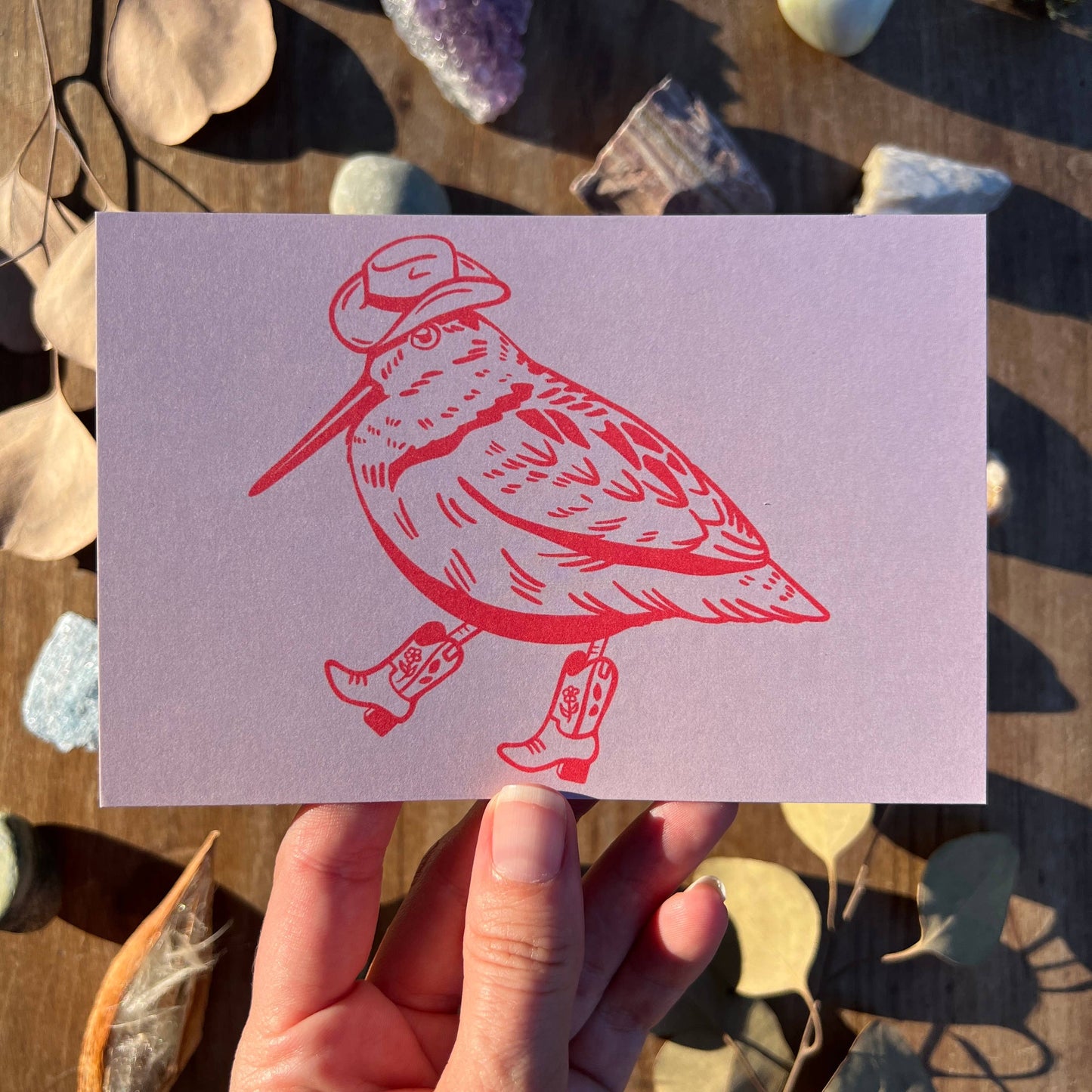 Card with a red bird illustration wearing boots and a hat held by a hand against a natural background.