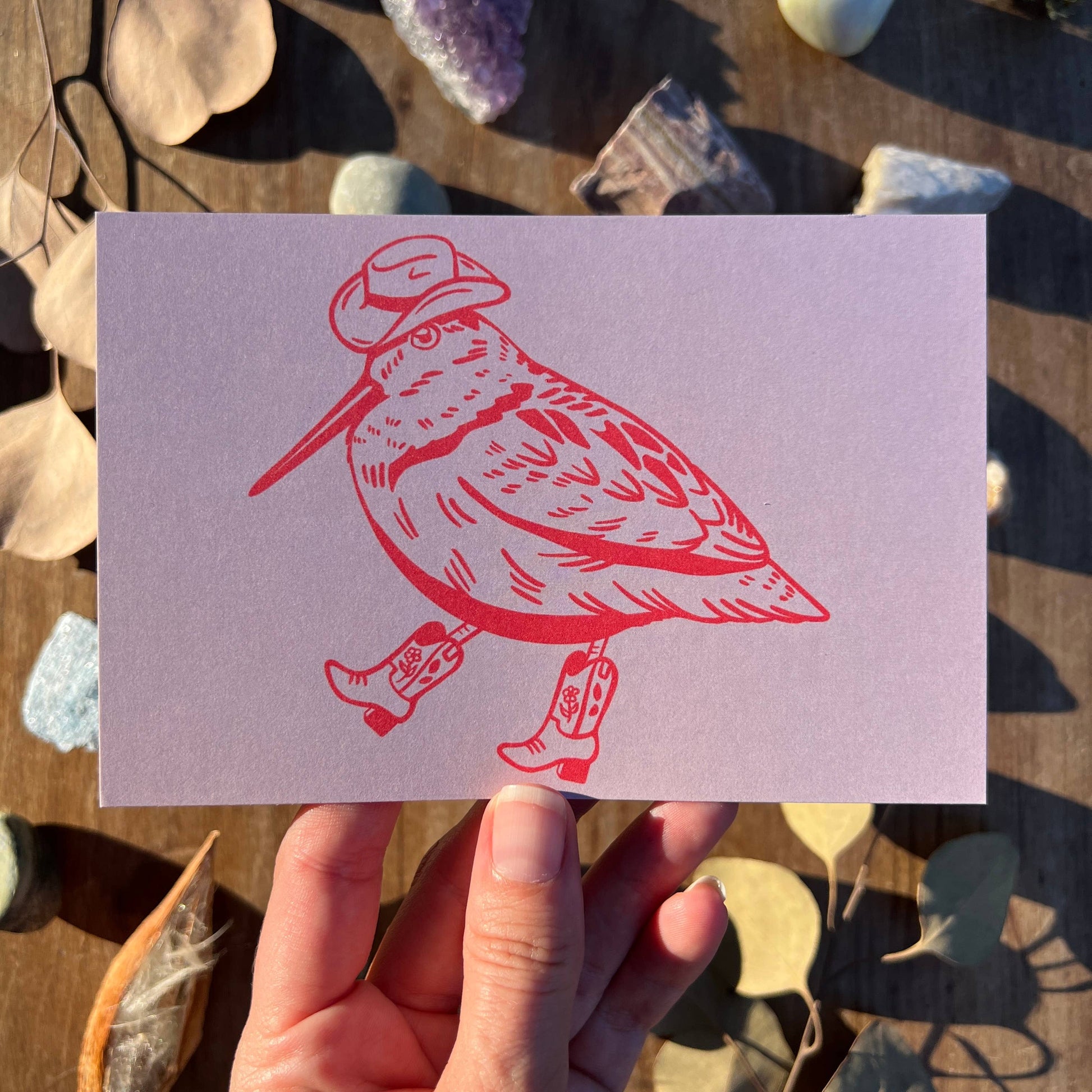 Card with a red bird illustration wearing boots and a hat held by a hand against a natural background.