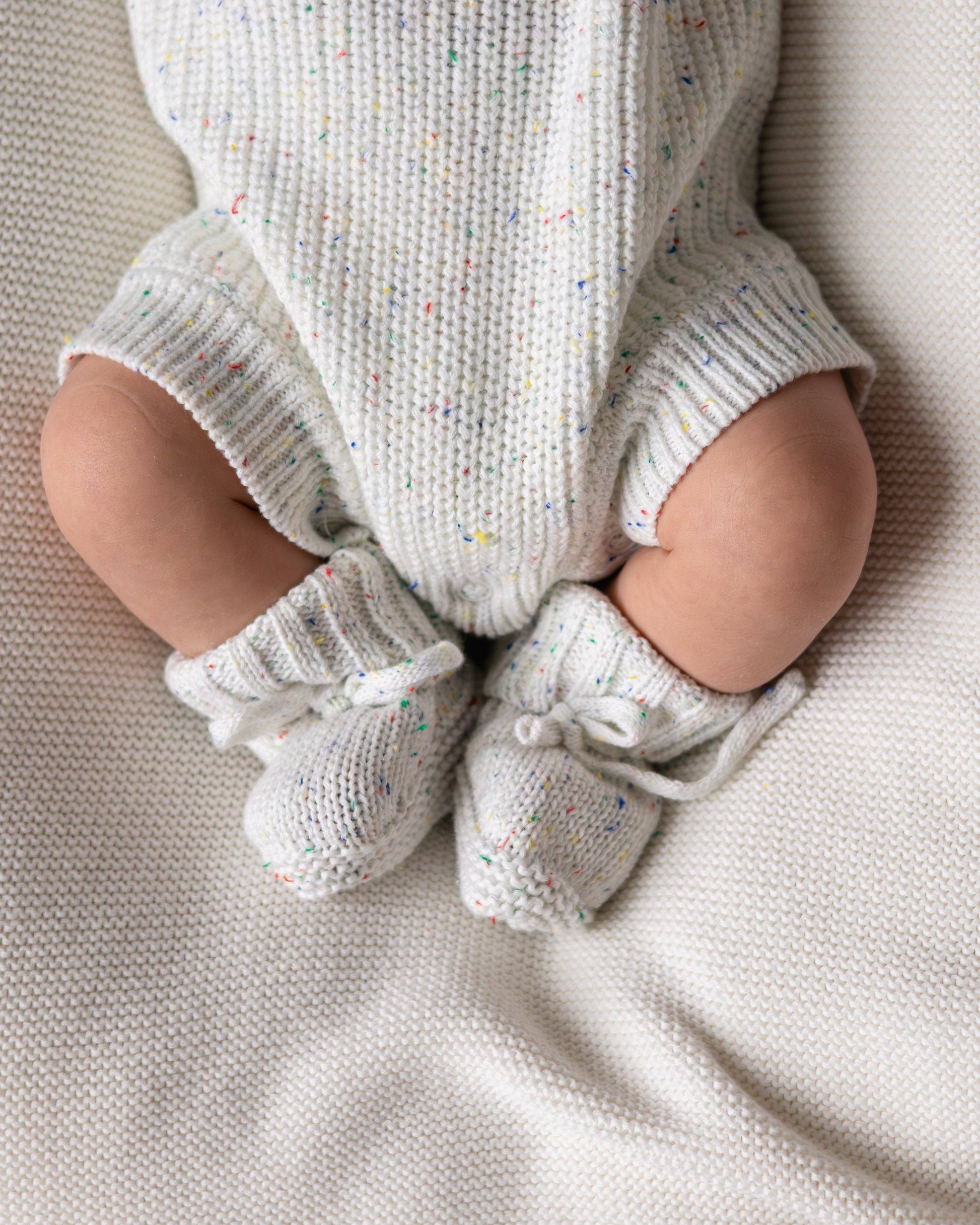 Baby legs wearing knitted gray romper and booties on a soft surface