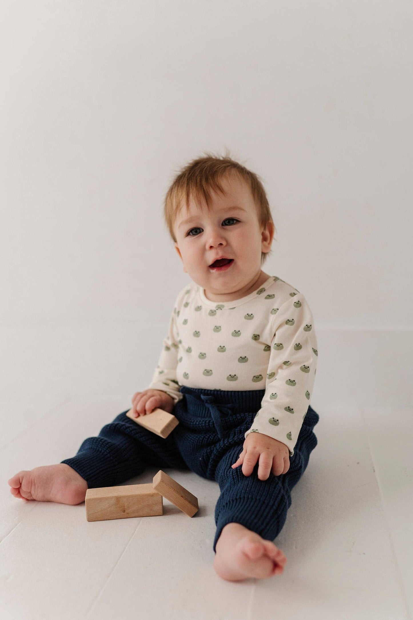 Baby sitting on a white floor with wooden blocks, wearing a patterned shirt and dark pants.