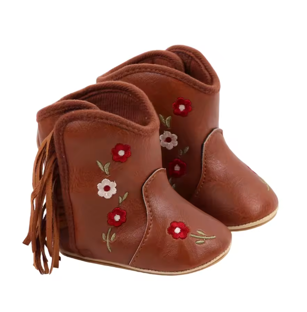 A pair of brown leather baby booties decorated with red and white floral embroidery and green leaf details, featuring a side fringe on one boot, set against a plain white background.