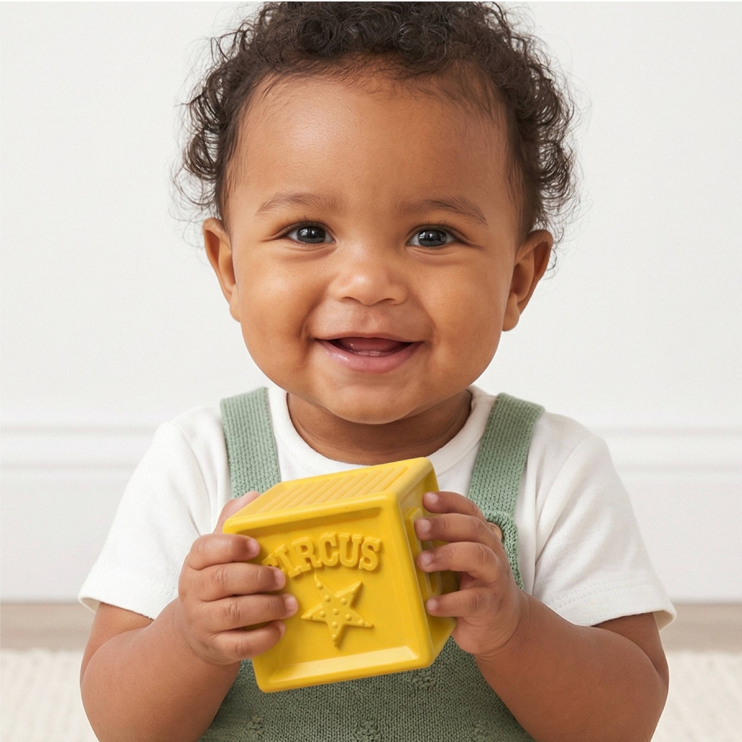 A smiling toddler in a white t-shirt and green overalls holds a bright yellow block toy embossed with the word 'CIRCUS' and a star. The child is photographed against a clean, off-white background.