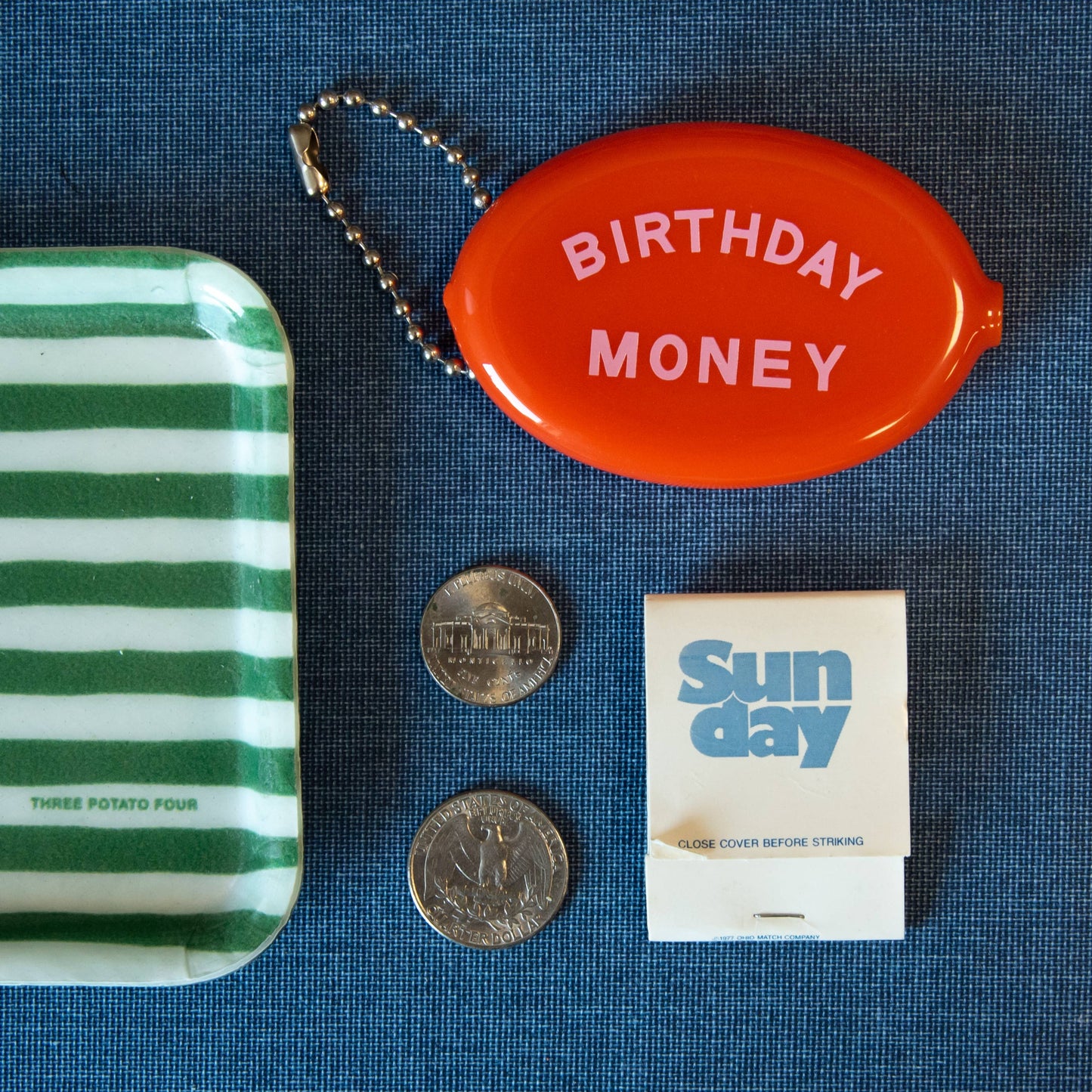 Green and white striped tray, red 'Birthday Money' container, coins, and a matchbook on a blue textured surface.