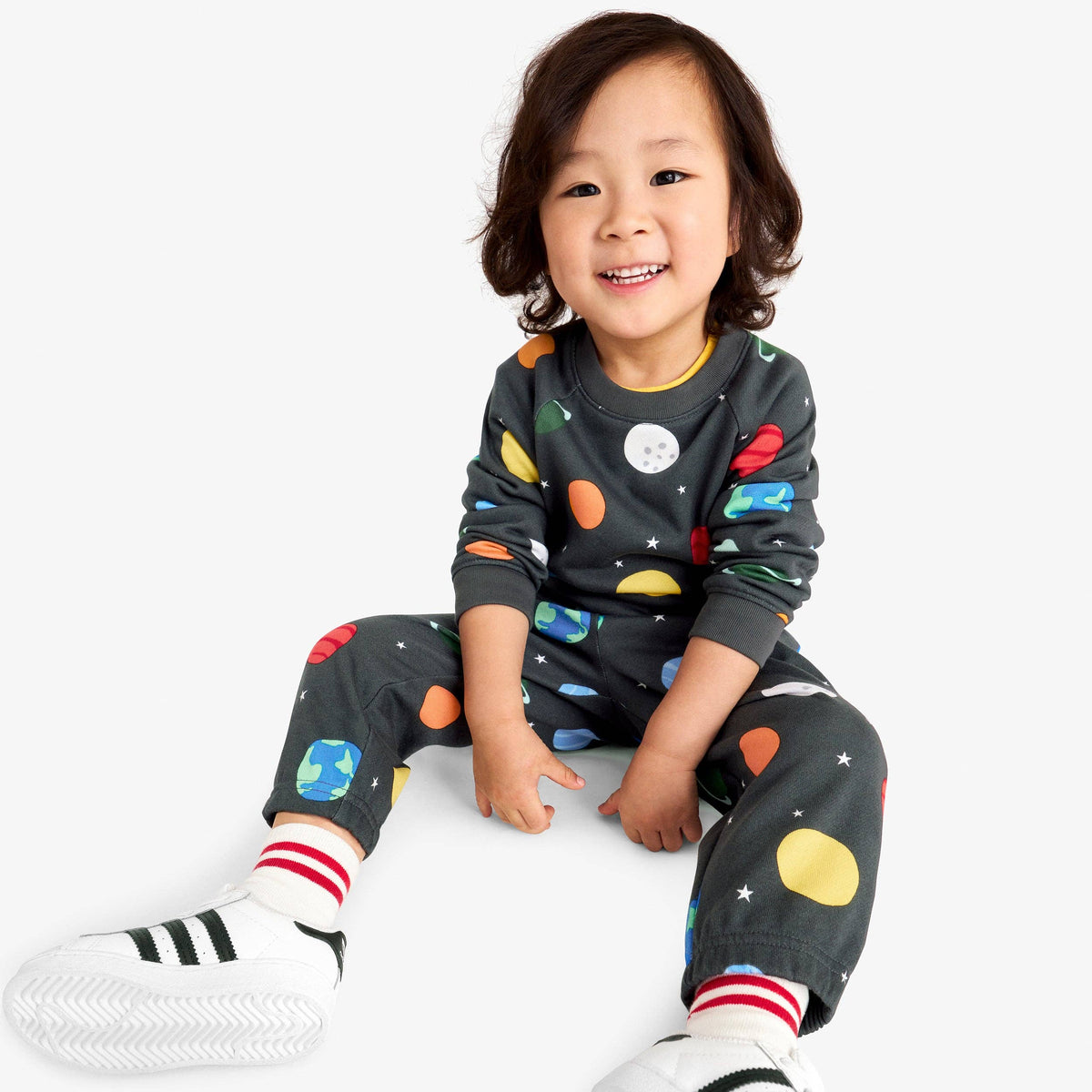 A smiling young child wearing a dark gray long-sleeve sweat set with a colorful space print, including planets, stars, and a moon button on the chest, paired with white sneakers and striped socks, sitting against a light gray background.