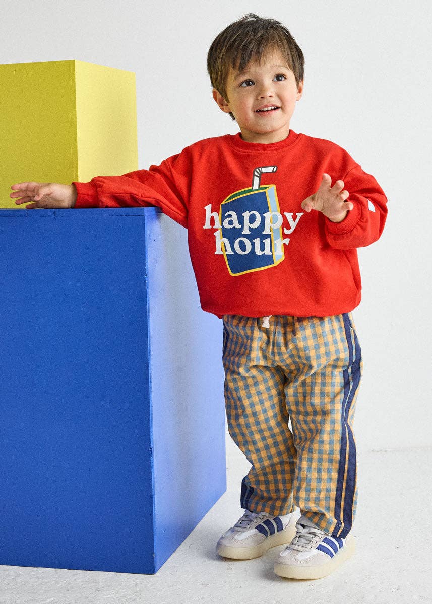A young boy wearing a red sweatshirt with a juice box graphic and the words 'happy hour' in white, paired with yellow and blue checkered pants, posing next to a blue and yellow block set against a white studio background.