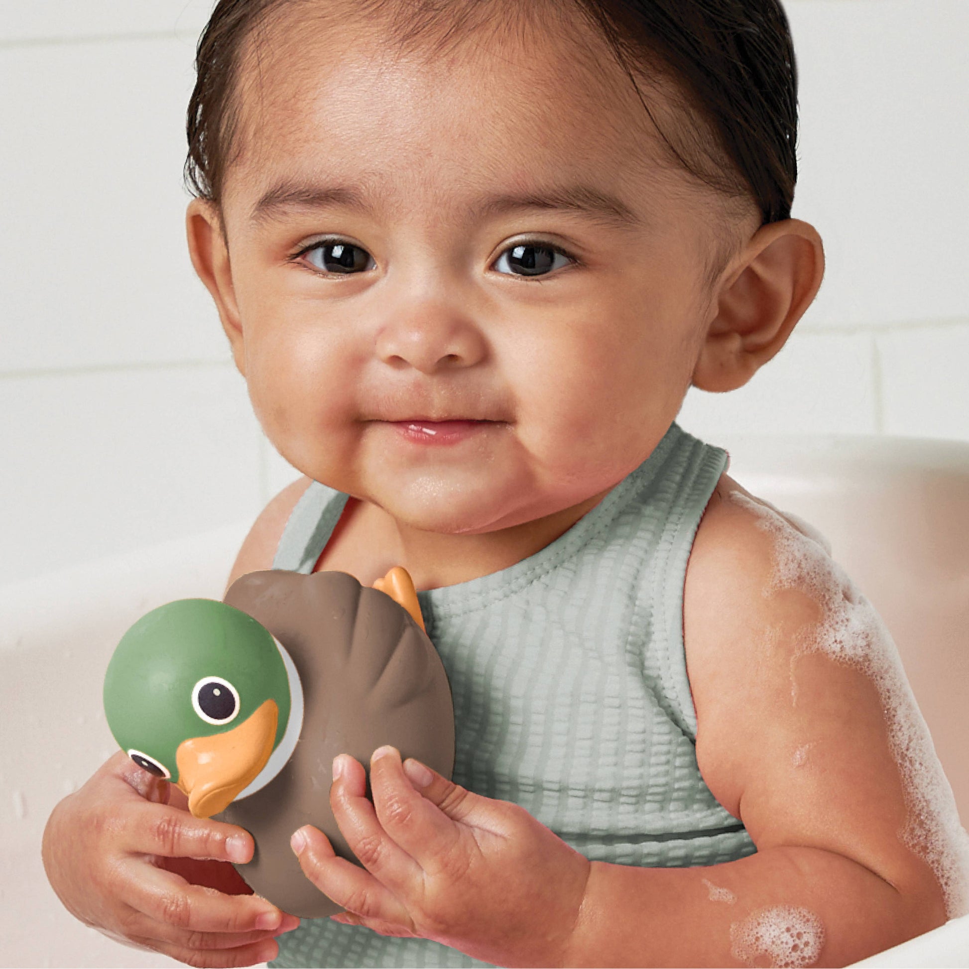 A baby in a light green waffle-knit onesie holds a duck-shaped bath toy with a green head, brown body, and orange beak while sitting in a white bathtub with soap foam on the skin.