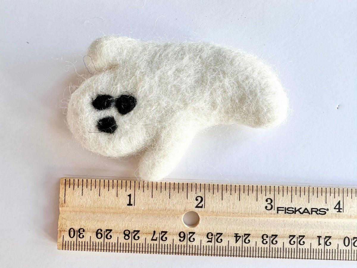 A small white felt ghost ornament with black eyes and mouth, placed above a wooden ruler on a white background.