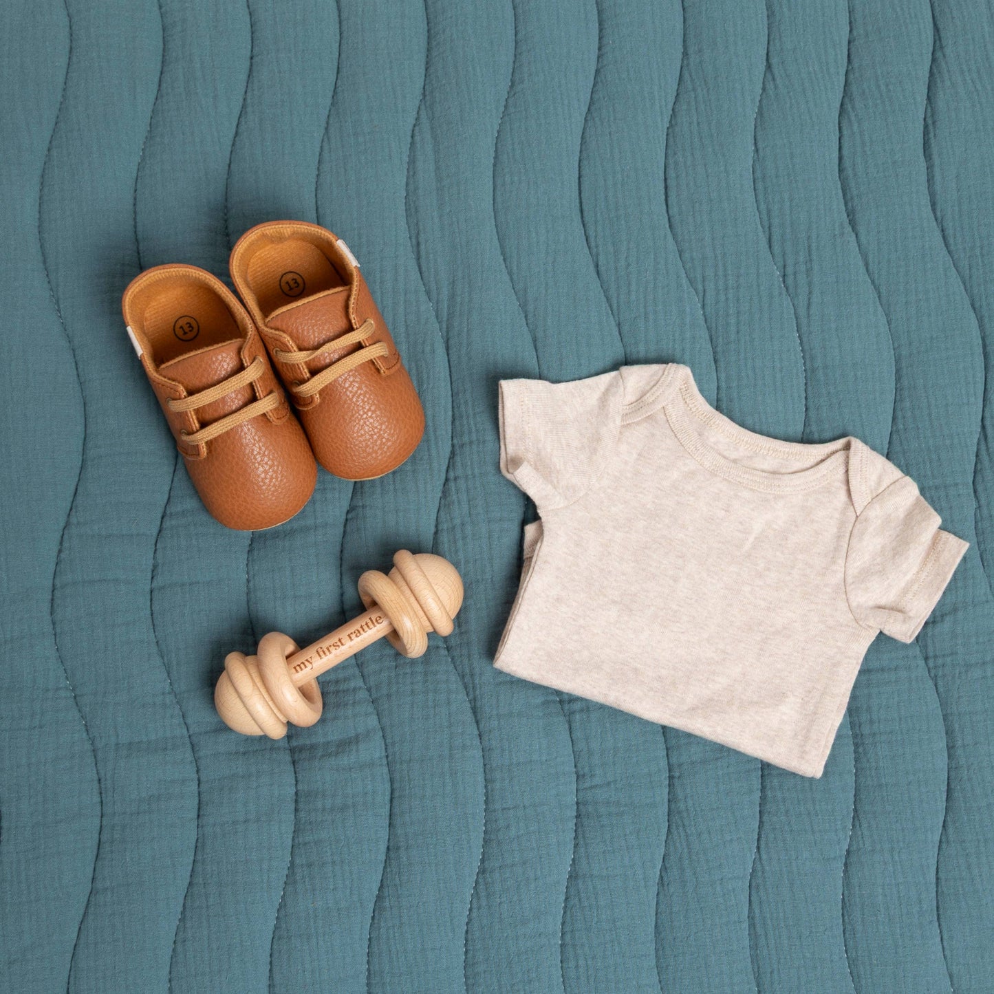 Pair of brown baby shoes, wooden rattle, and white baby shirt on a textured teal surface