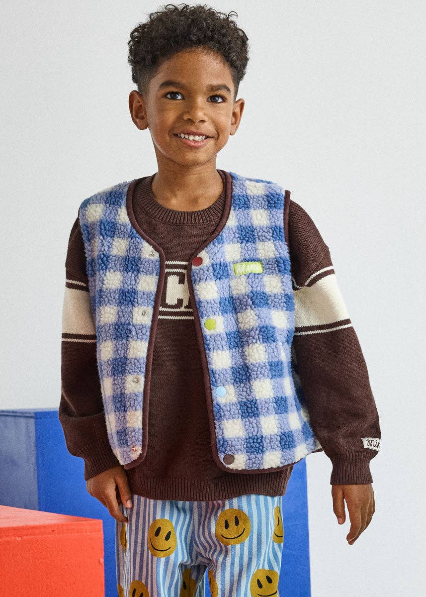 A young boy smiling and wearing a blue and white checkered fleece vest over a brown knit sweater, paired with blue striped pants featuring a yellow smiley face pattern.