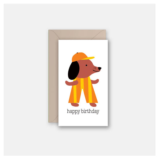A birthday greeting card with a white front featuring a cartoon dog in an orange and yellow striped outfit and cap, with the text 'happy birthday' below. The card is shown with a plain brown envelope behind it on a light gray background.