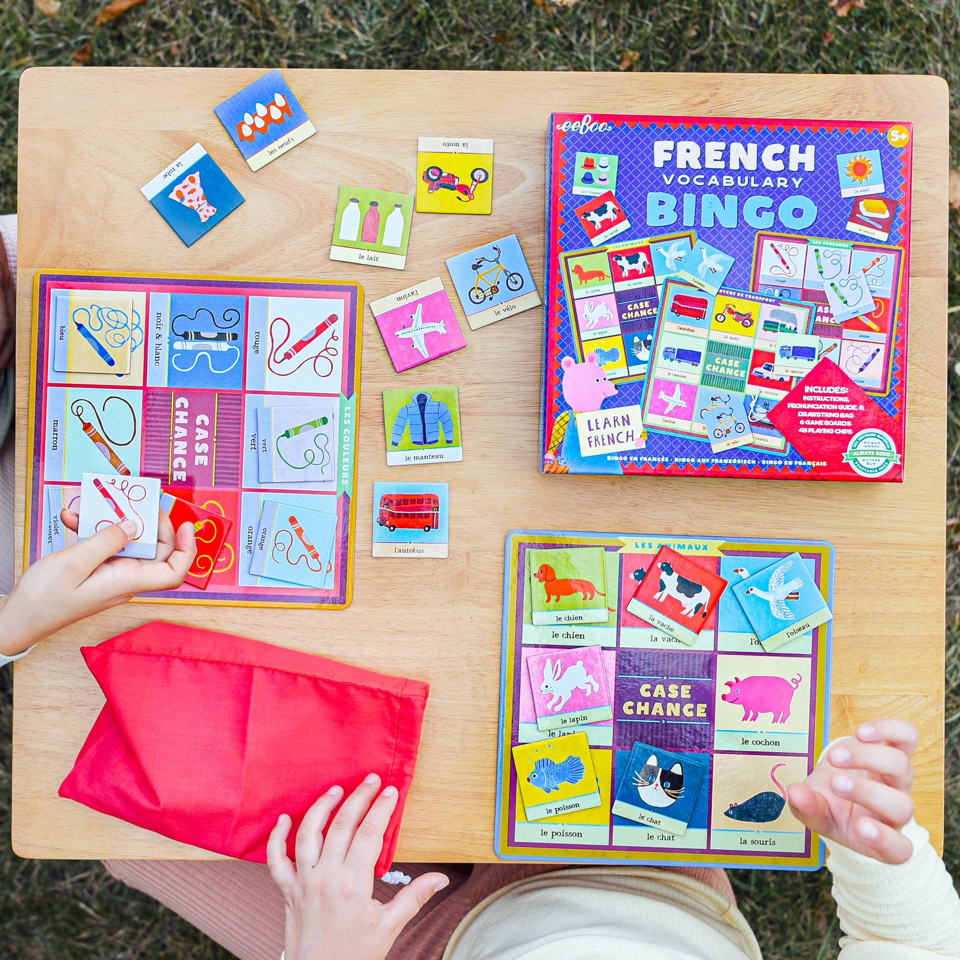 Children playing with French vocabulary bingo game on a wooden table outdoors.