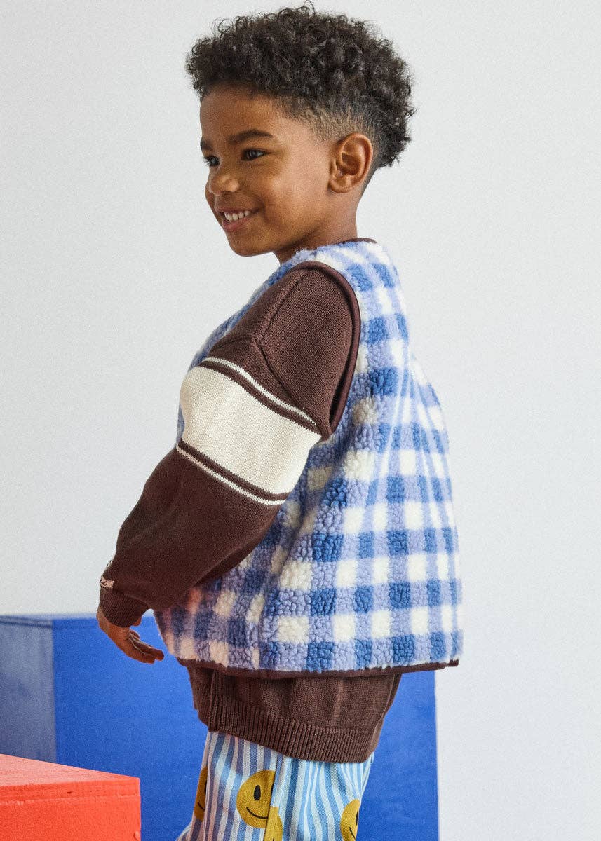 A young child wearing a blue and white gingham patterned vest over a brown and cream striped sweater, paired with blue striped pants featuring yellow smiley faces, posed against a light gray wall with colorful blocks.
