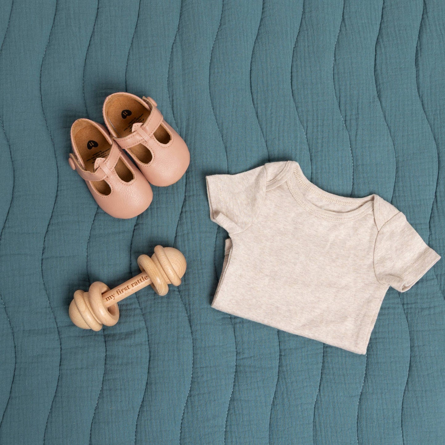 Baby shoes, wooden rattle, and t-shirt on a textured teal surface