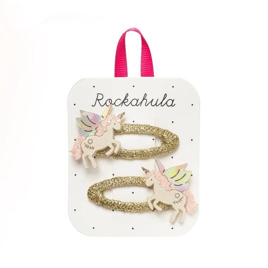 Two gold glitter unicorn hair clips with pastel wings and cream bodies, attached to a white display card with a pink ribbon loop. The card is branded 'Rockahula'.