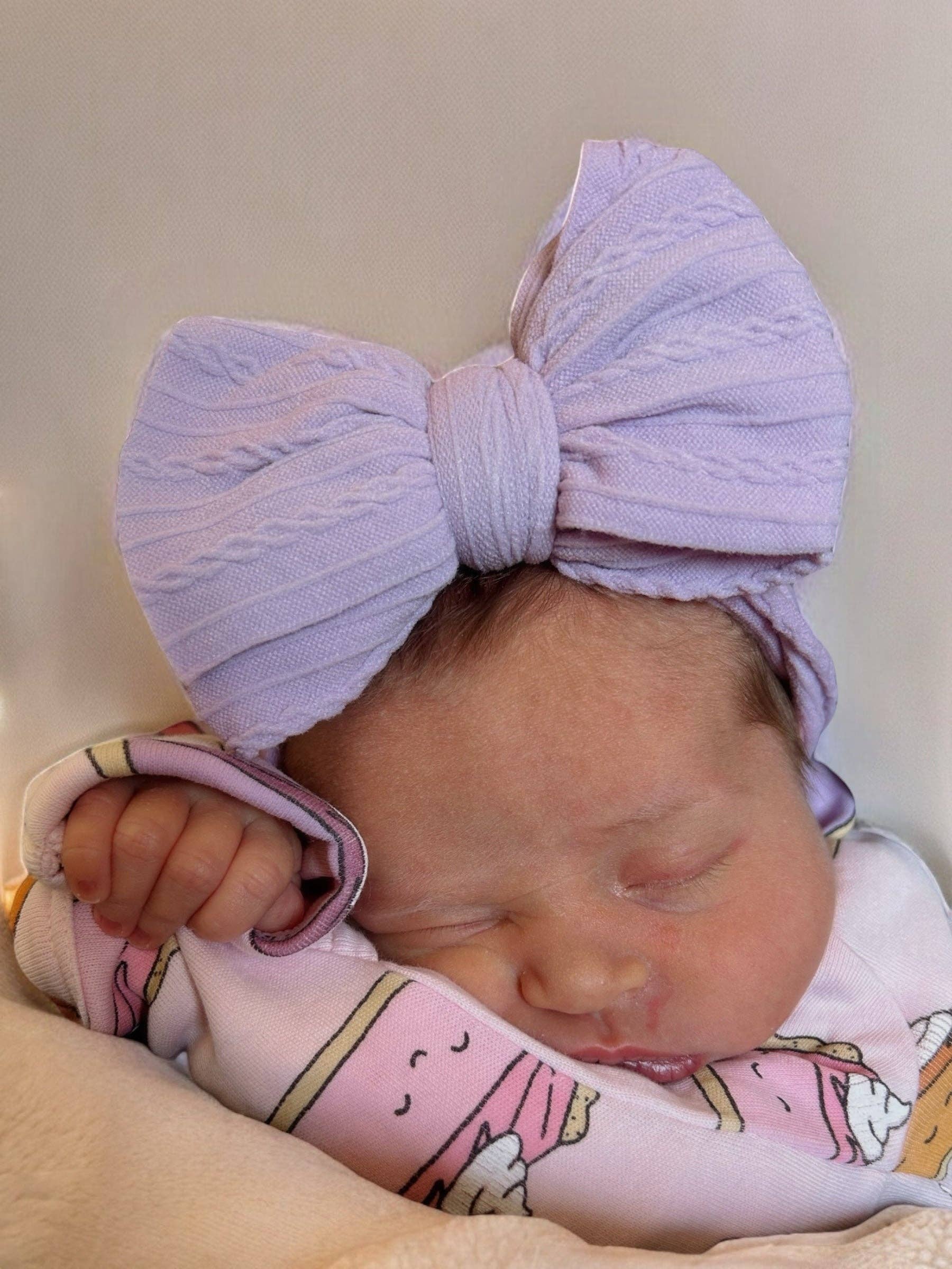 Newborn baby sleeping with a large purple bow on its head and a pink blanket.