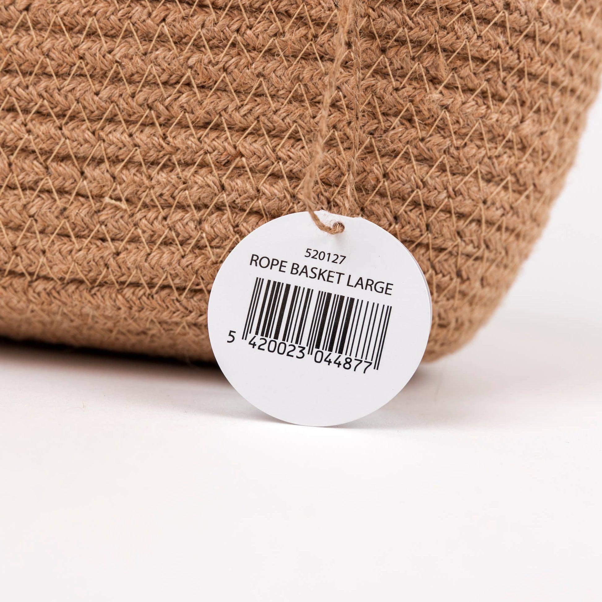 A close-up of a woven rope basket in natural tan jute with a white circular product tag reading 'ROPE BASKET LARGE' and a barcode, photographed on a white background.