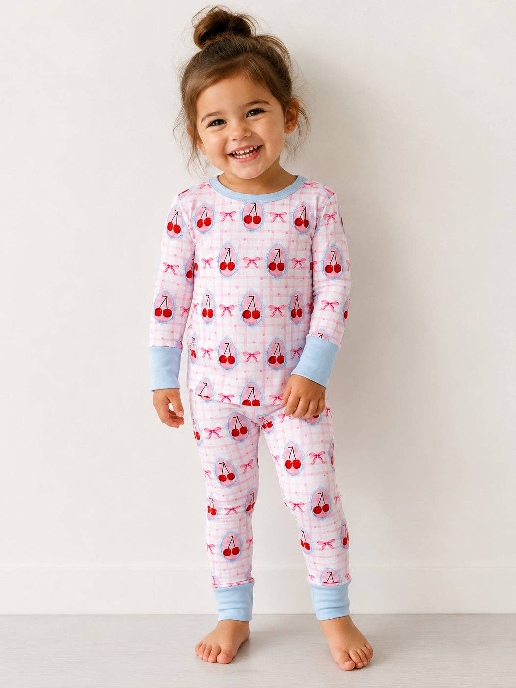 A young girl smiling and wearing a long-sleeve pajama set with a cherry print. The pajamas are light pink with a subtle grid pattern, decorated with red cherries and pink bows, and finished with light blue cuffs and collar.