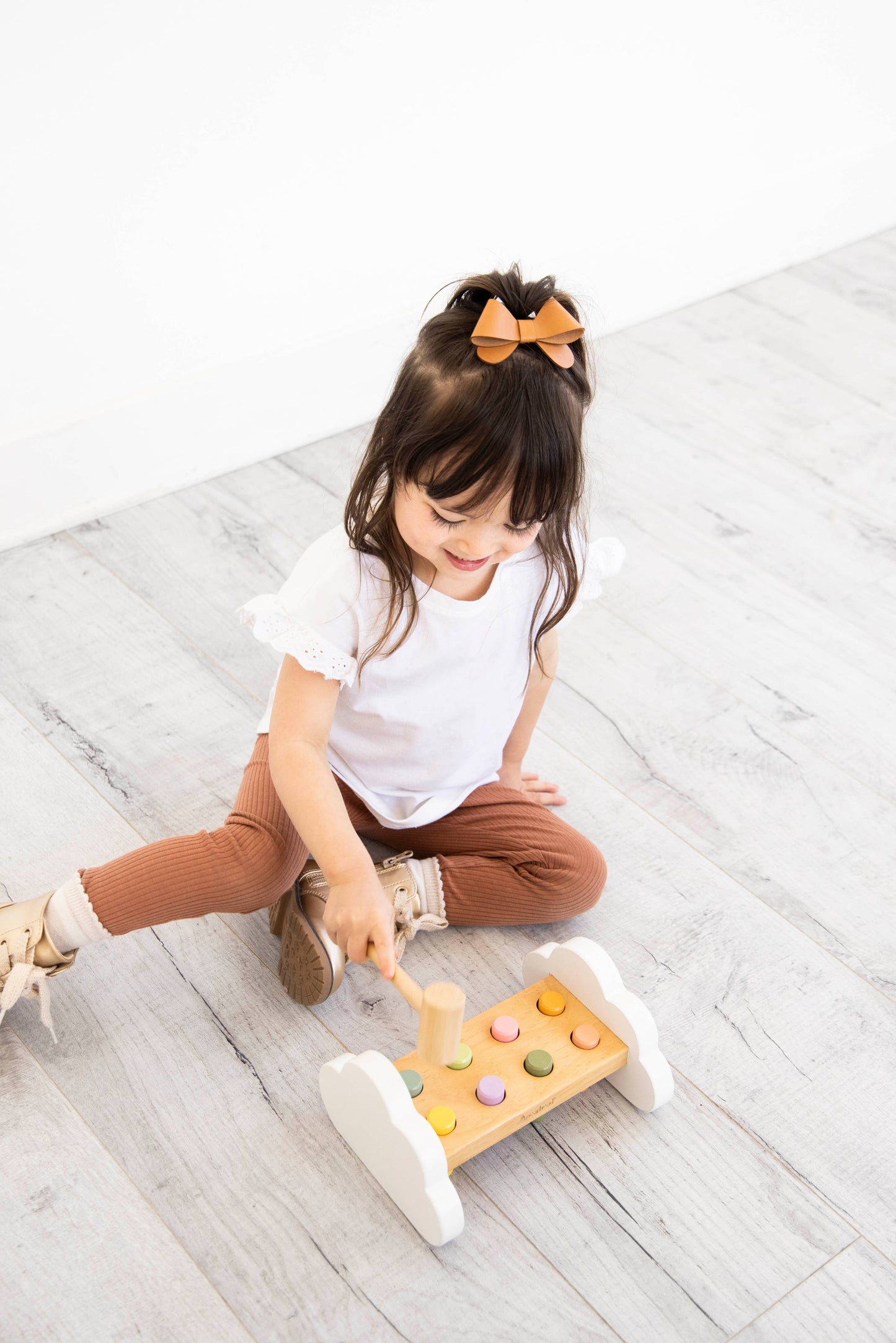 A young child sits on a wooden floor in front of a white wall playing with a wooden hammer bench toy with colorful pegs and a wooden mallet.